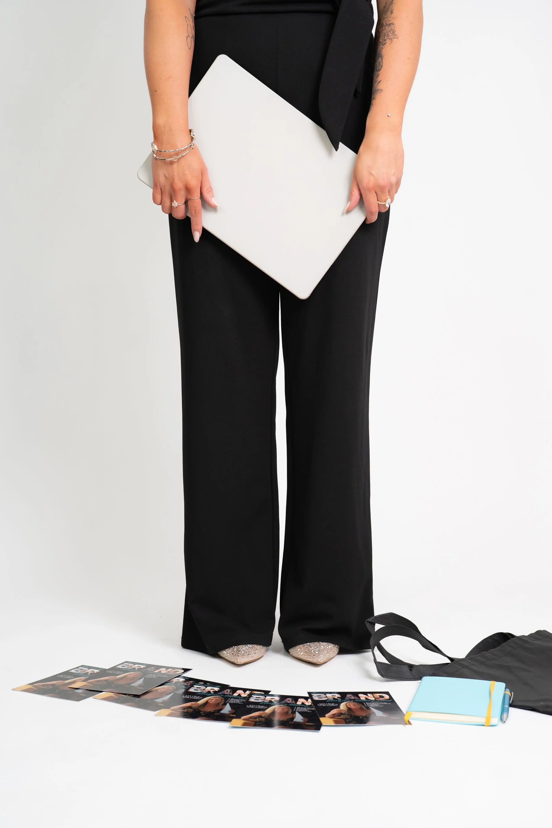 Person wearing black pants and glittery shoes holding a closed white laptop, surrounded by magazines, a notebook, a backpack, and a water bottle on the floor.