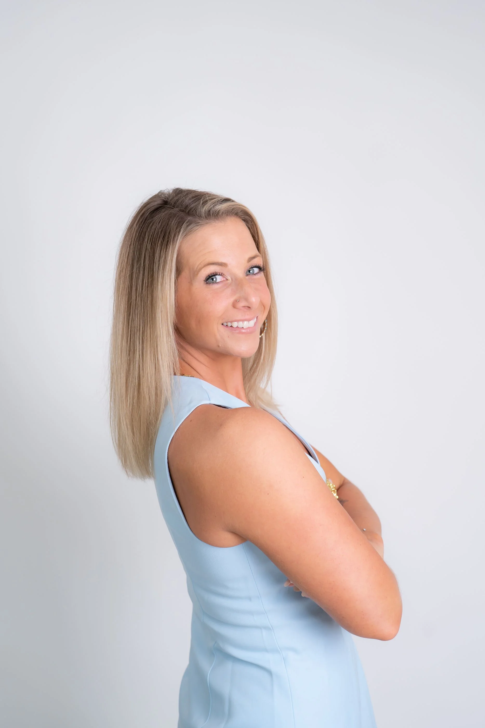 Smiling woman with blonde hair in a light blue sleeveless dress, posing with arms crossed against a plain white background.