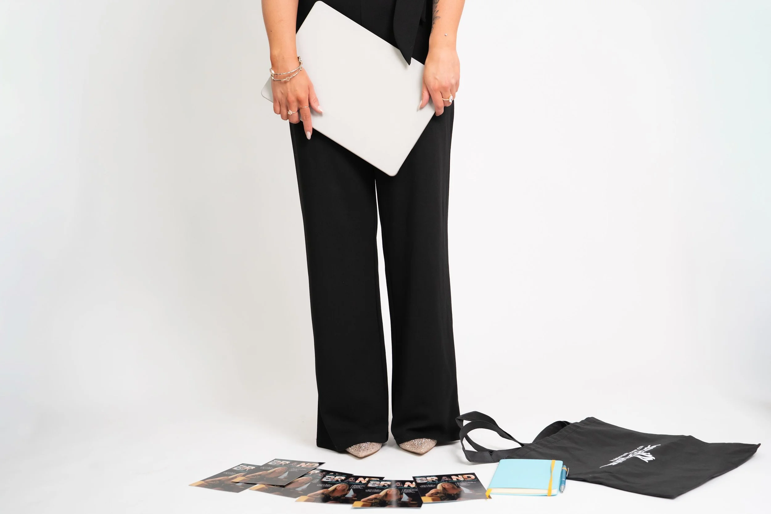 Person holding a white folder or book, wearing black pants and jewelry, standing on a white surface with various magazines, a notebook, and a black tote bag on the floor.