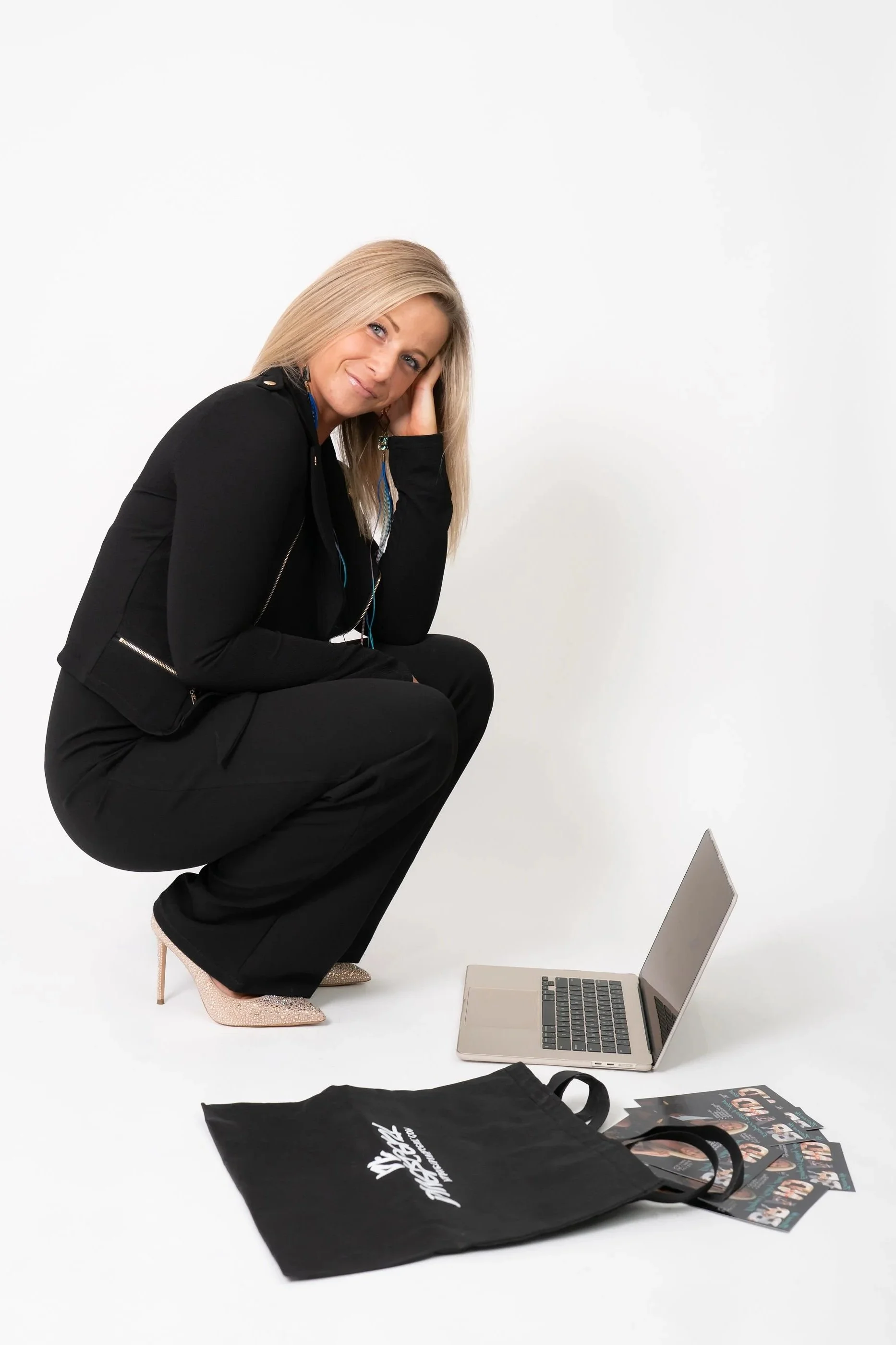 A woman in black clothing and sparkly high heels squats in front of a laptop, holding her head with one hand, with a tote bag and printed materials on the ground, against a plain white background.