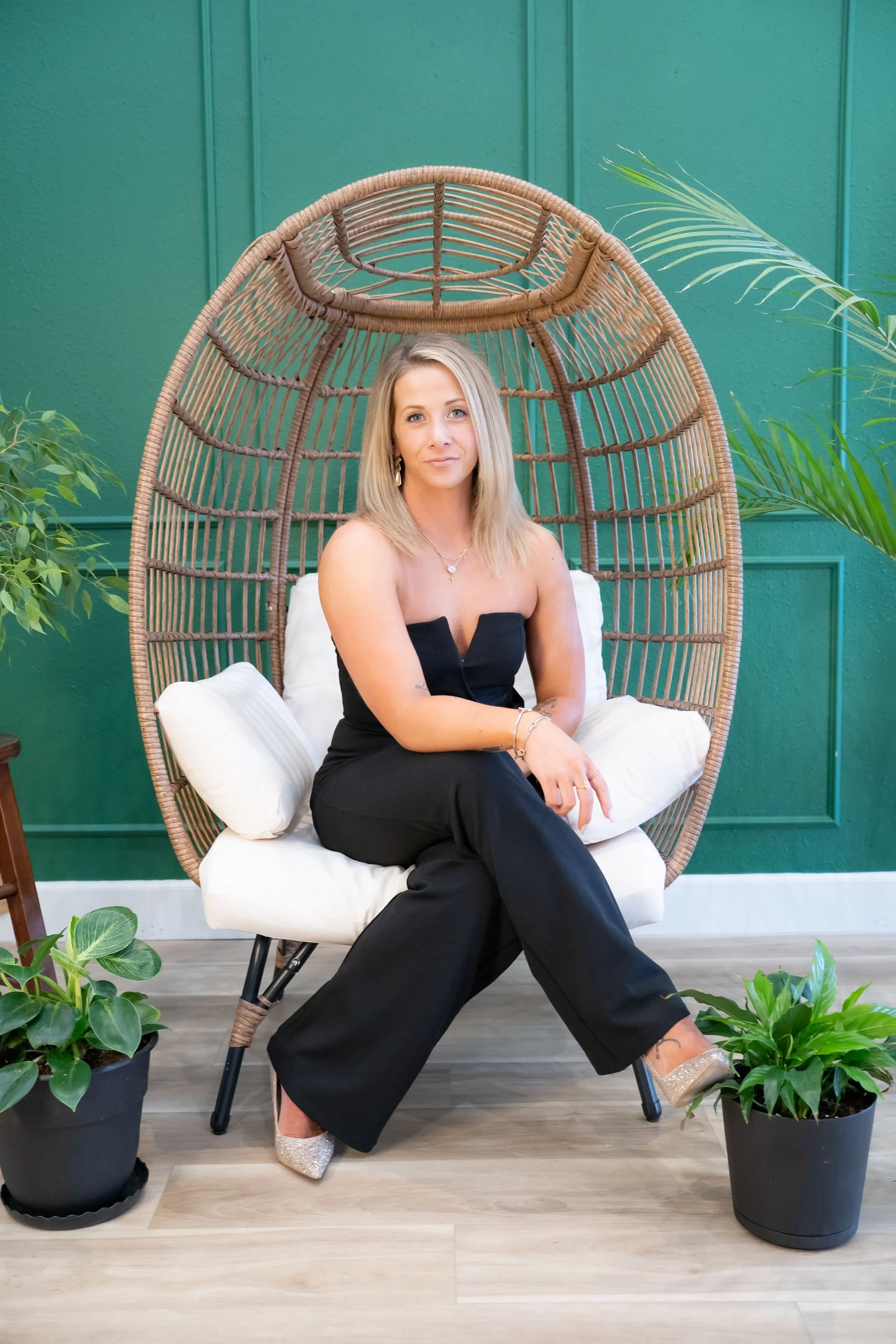 A woman with blonde hair wearing a black strapless jumpsuit, sitting on a white cushion in a rattan hanging chair, surrounded by green plants, against a teal wall.