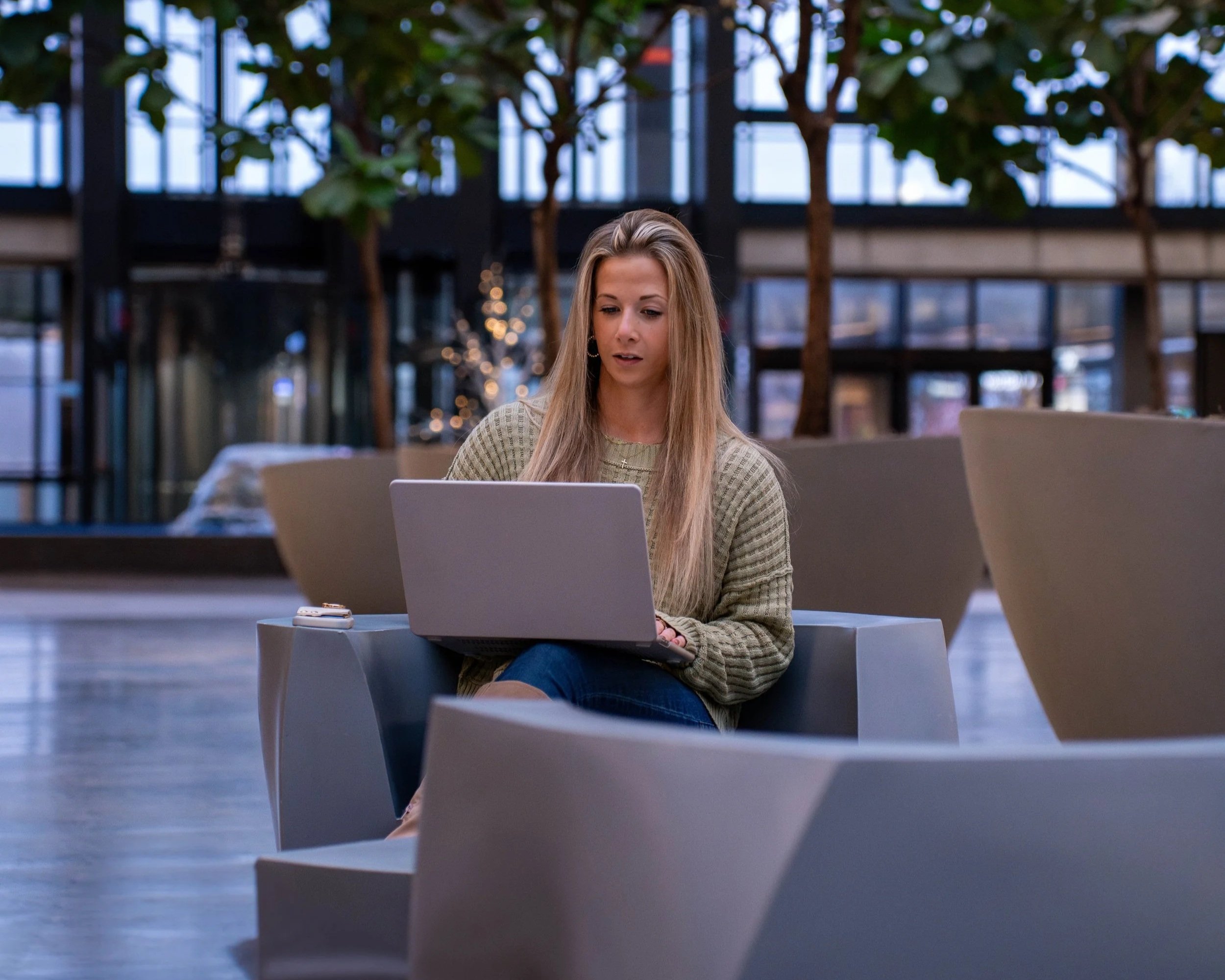 A woman with long blonde hair sitting on a modern indoor chair with a laptop on her lap, in a spacious, well-lit indoor area with large windows and indoor trees.