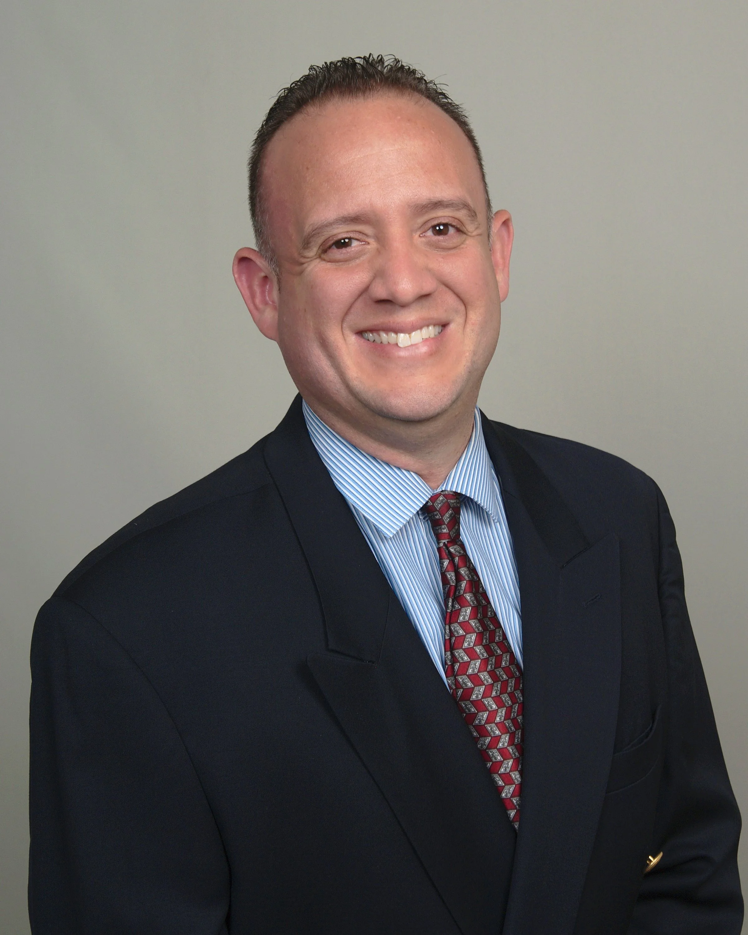 A smiling man in a dark suit, blue striped shirt, and red patterned tie against a plain background.