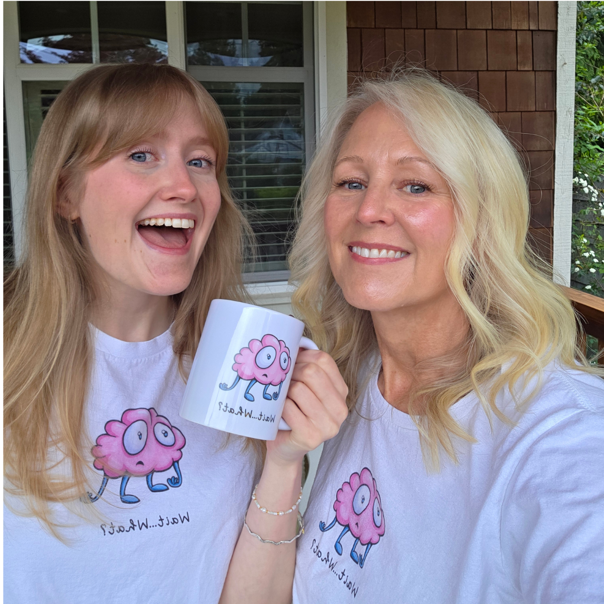A smiling young woman and an older woman, both wearing matching white T-shirts with a pink brain character and the words 'Well...What?' They are outside near a house with a window behind them, holding a white mug with the same pink brain character, and both appear happy.