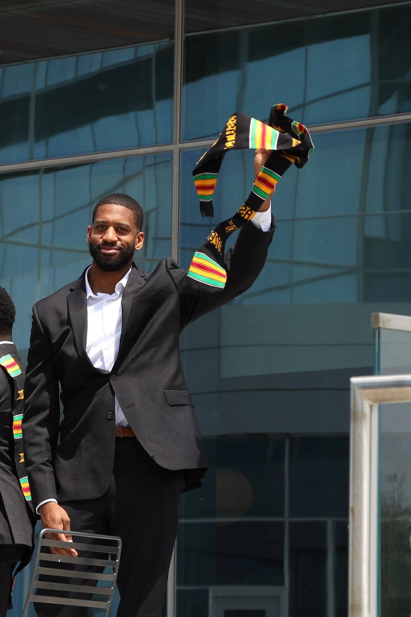 A man in a suit smiling and holding a Kente cloth, standing outside of a glass building.