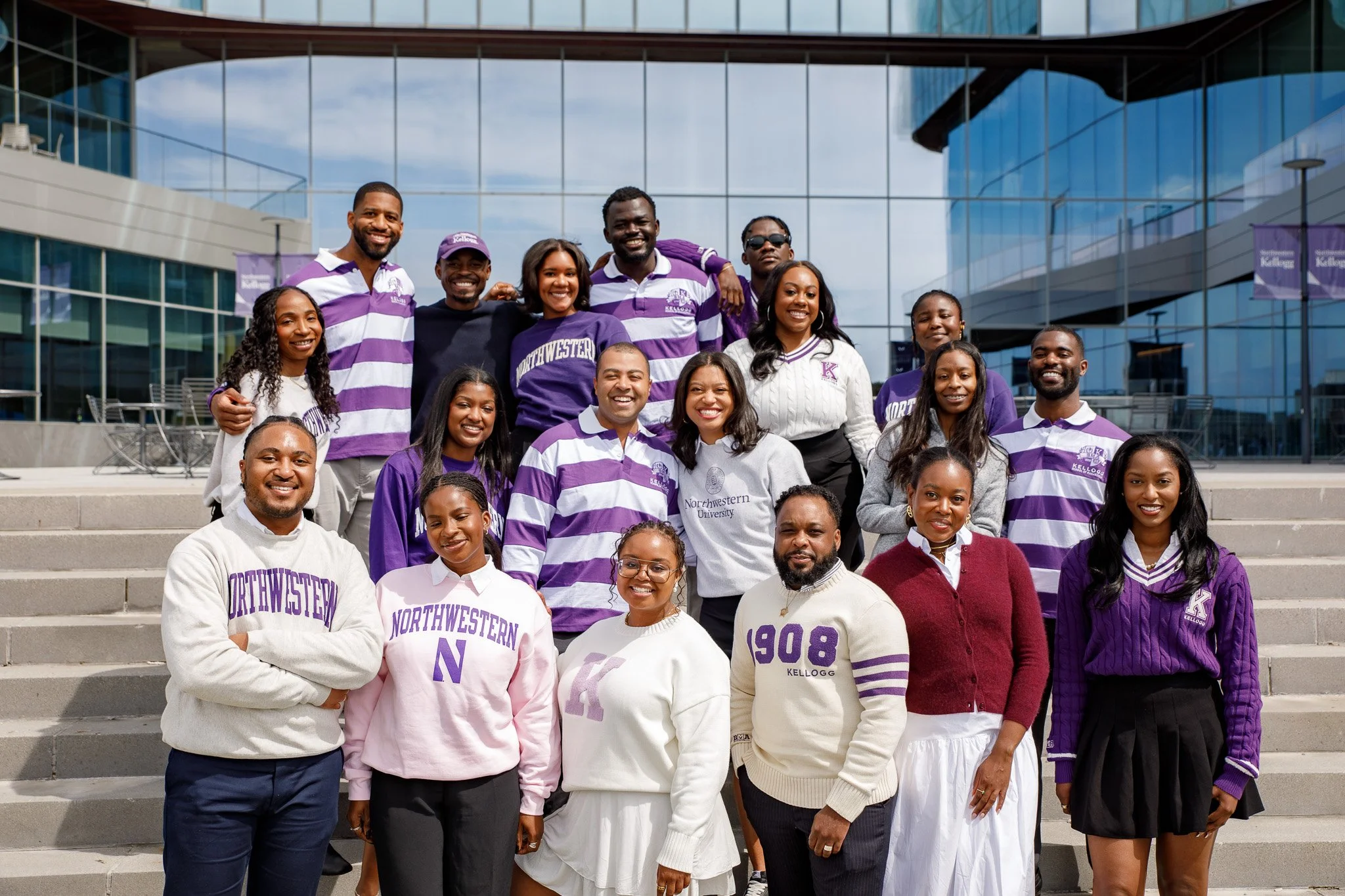 Group of diverse university students standing on steps outside modern glass building, wearing Northwestern University clothing, smiling, with some making peace signs.