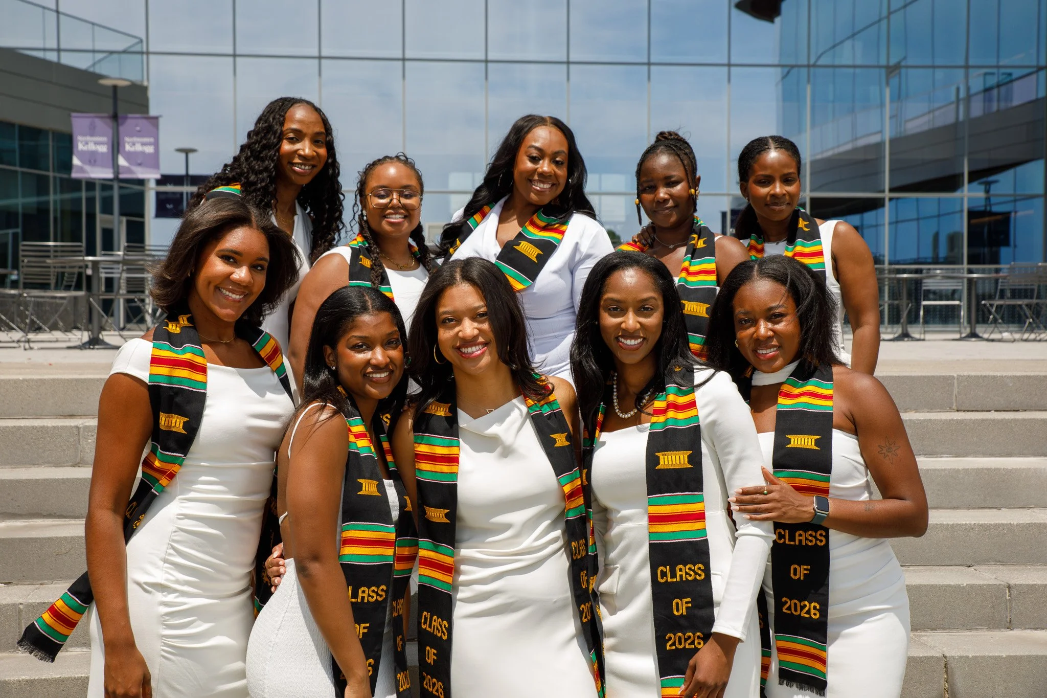 A group of diverse young women dressed in white gowns and stoles with colorful patterns celebrating graduation outside a modern glass building.
