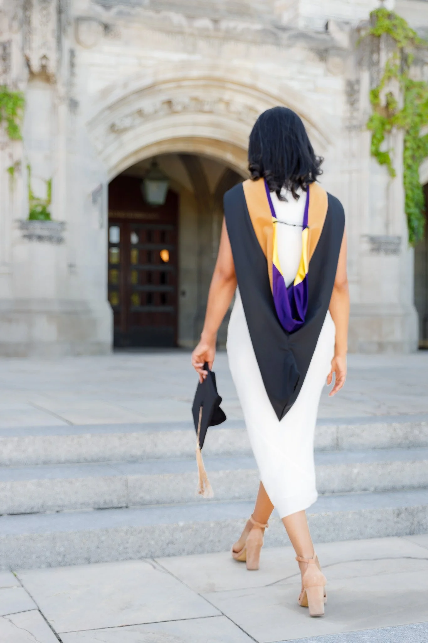 Woman in a white dress with a large black and yellow scarf walking up steps in front of an ornate stone building.