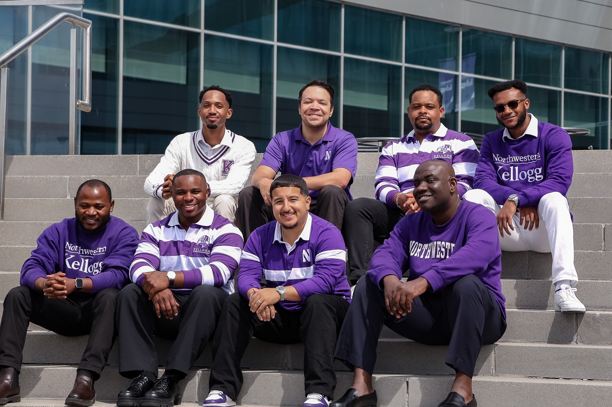 Group of nine diverse college students sitting on steps outside modern glass building, wearing purple and white University of Northwestern Kellogg shirts and sweaters, smiling at the camera.
