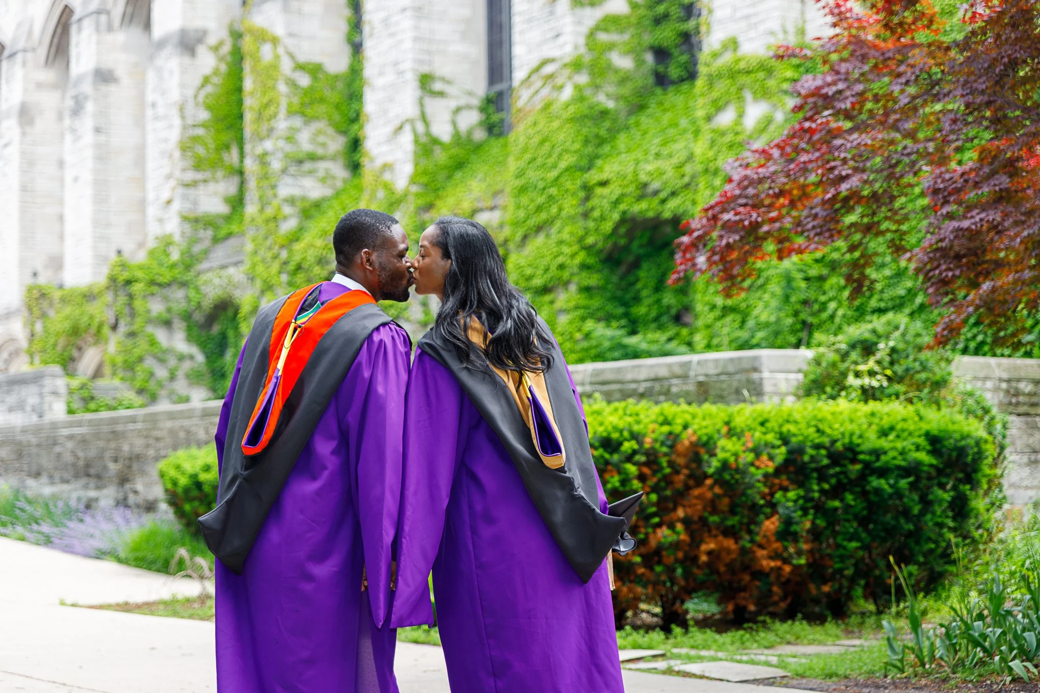 Two graduates in purple caps and gowns sharing a kiss outdoors during graduation ceremony surrounded by lush greenery and blooming plants.