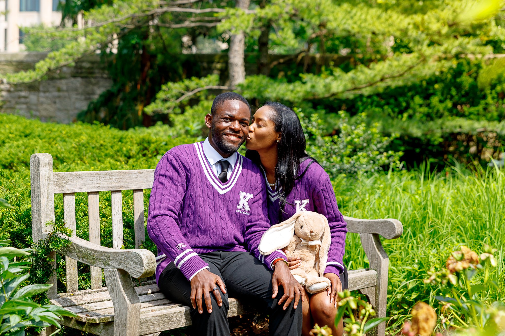 A couple sitting on a wooden park bench, dressed in purple sweaters with Greek letters, surrounded by green trees and bushes, with the woman kissing the man's cheek.