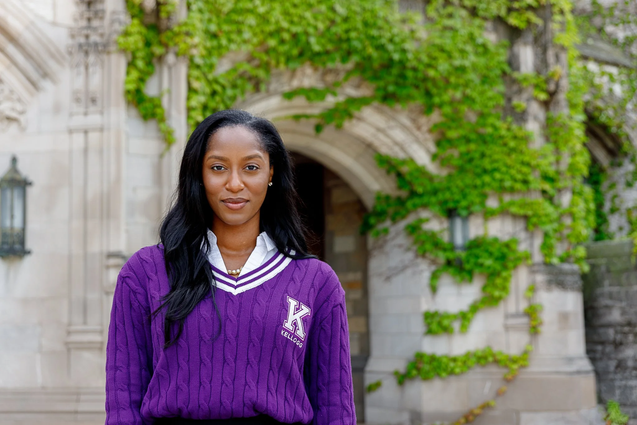A woman with long black hair wearing a purple sweater with Greek letters, standing outdoors in front of a building covered in green ivy.