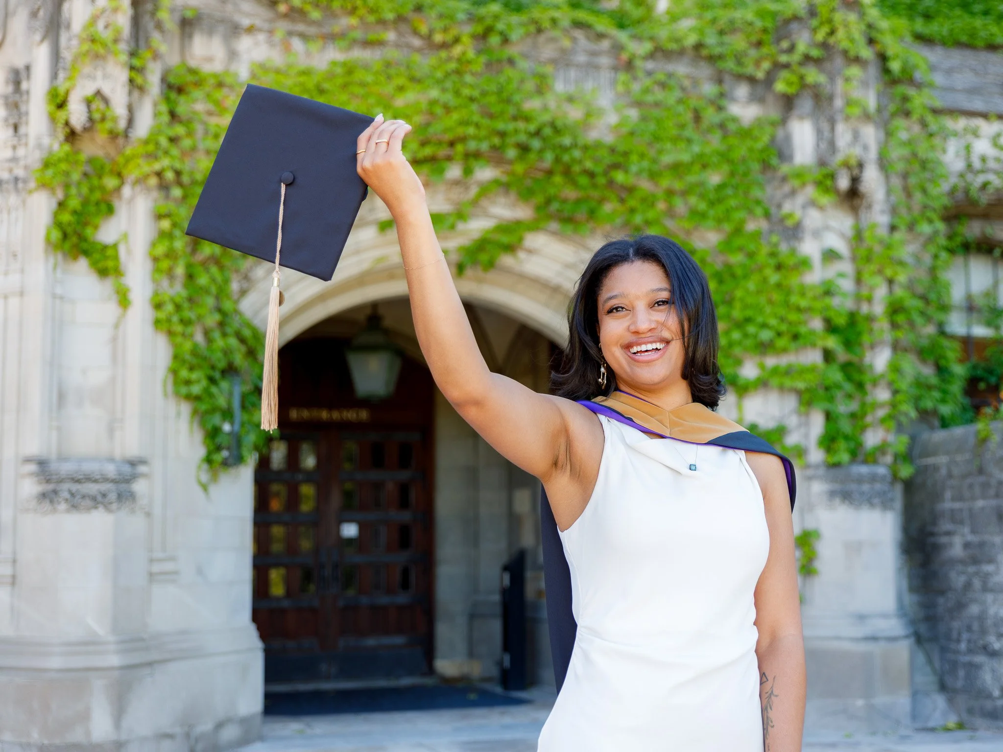 A young woman with dark hair, smiling and wearing a graduation cap and gown, holding the cap up in the air outdoors with green ivy and a stone building in the background.