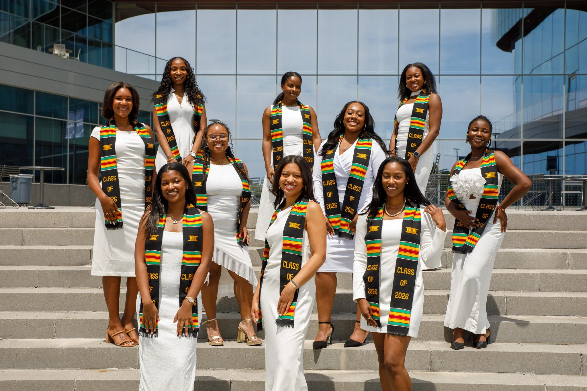 Group of women in white dresses celebrating graduation outdoors, wearing black stoles with colorful stripes and gold text that reads "Class of 2026" in front of a modern glass building.