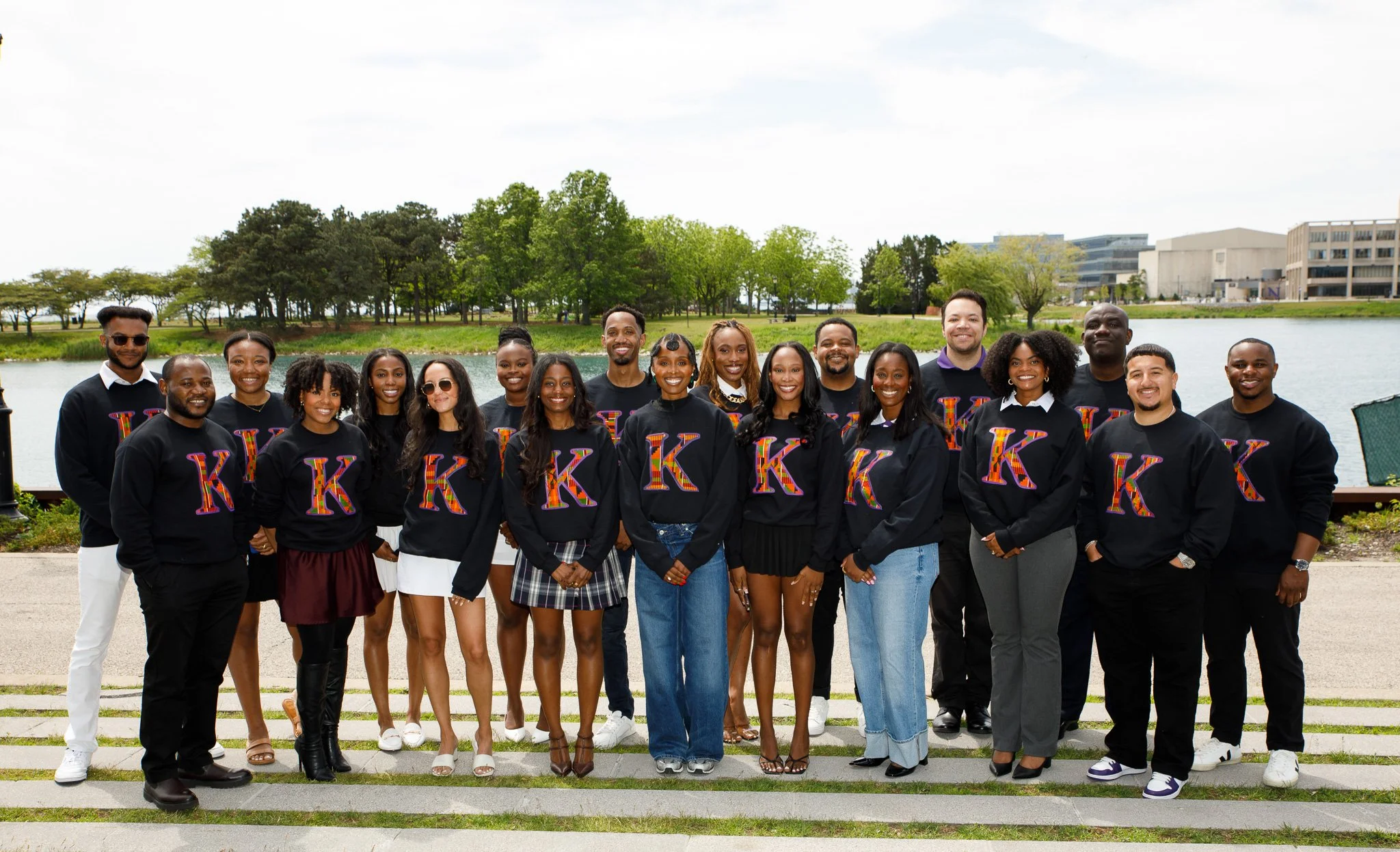 A diverse group of young adults posing outdoors near a body of water, wearing matching black sweatshirts with colorful Greek letters, with trees and buildings in the background.