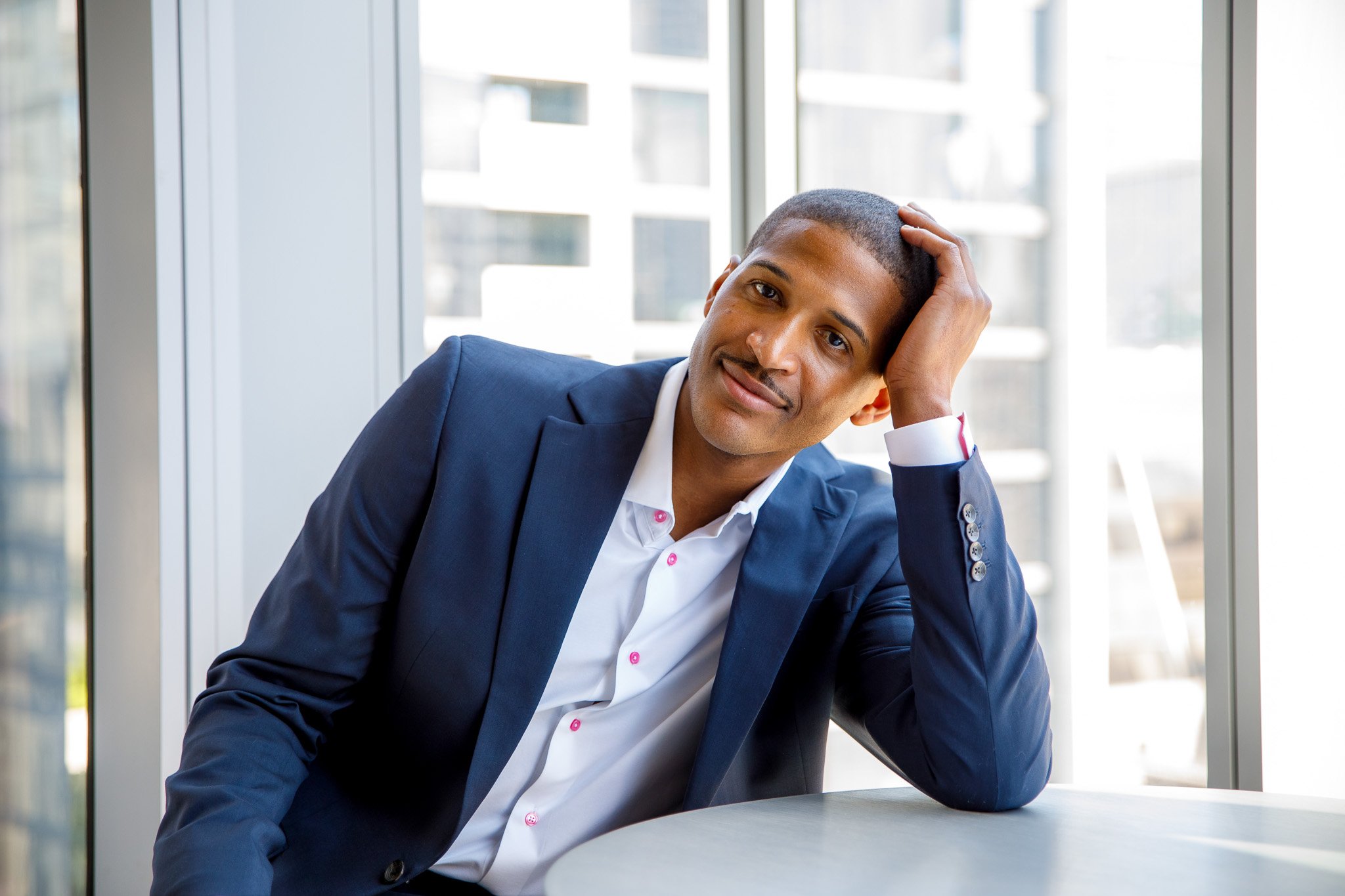 A young man in a blue suit and white shirt sitting at a table near large windows, resting his head on his hand and looking thoughtfully to the side.