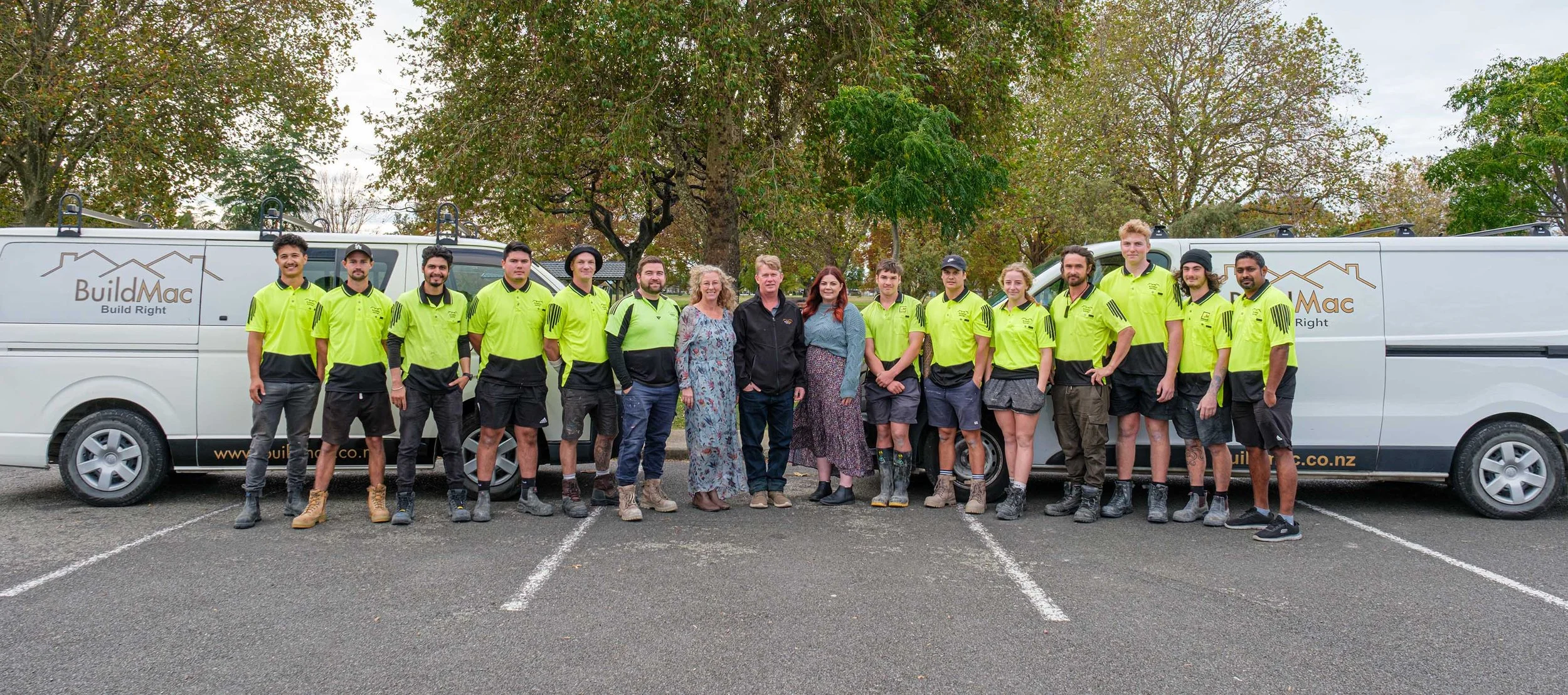Group of construction workers and staff standing in front of two company vans with trees in the background. Most are wearing bright yellow shirts, some are in casual clothing.