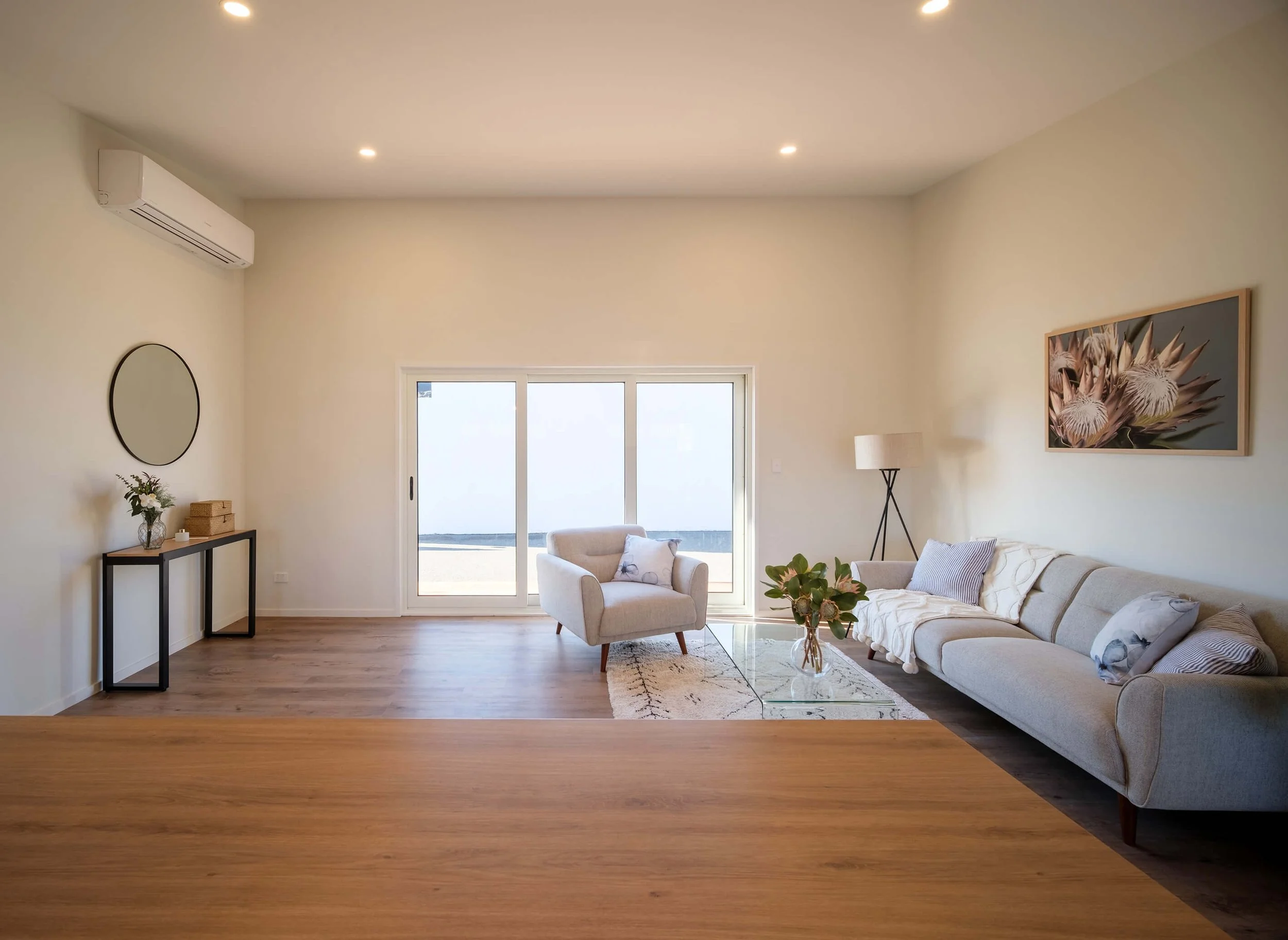 Living room with white walls, hardwood floors,  a glass coffee table with a plant, a beige armchair, a floor lamp, a corner artwork of flowers, and a large sliding door leading outside. Perfect for enjoying the Hawkes bay sun