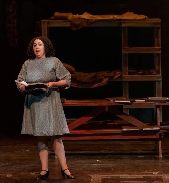Cornelia performing on stage in a staged production of Lori Laitman's Vedem, holding a journal, standing in front of wooden furniture and a background of shelves with folded fabric.