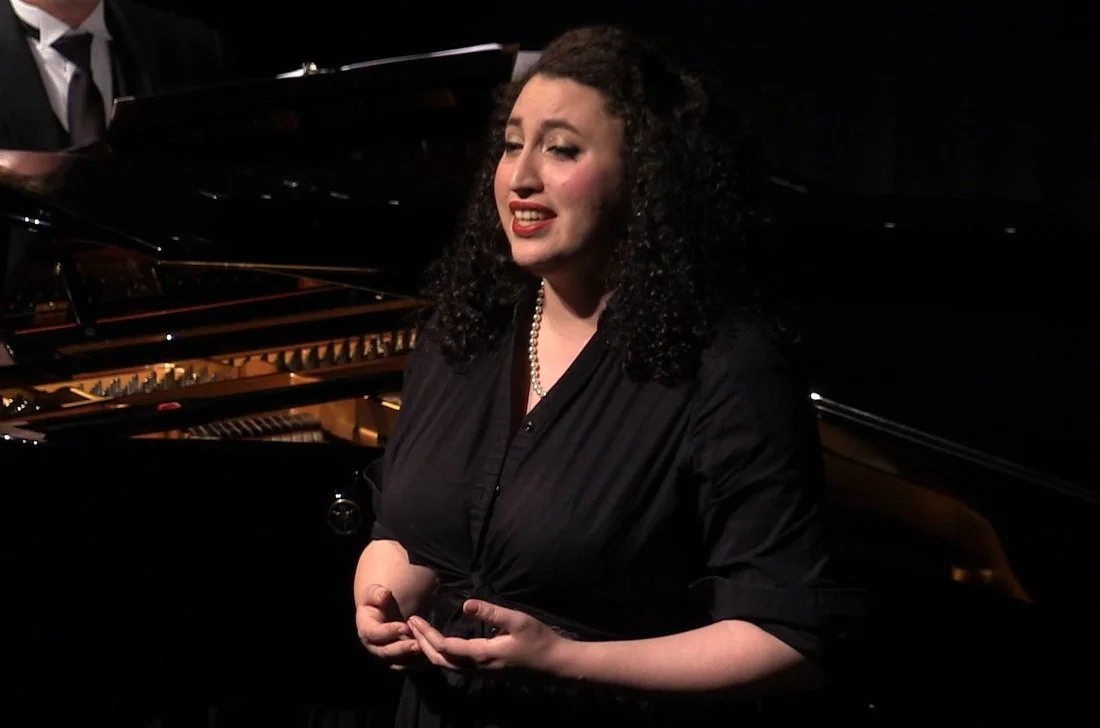 Woman singing on stage with a piano in the background, wearing a black dress and pearl necklace.