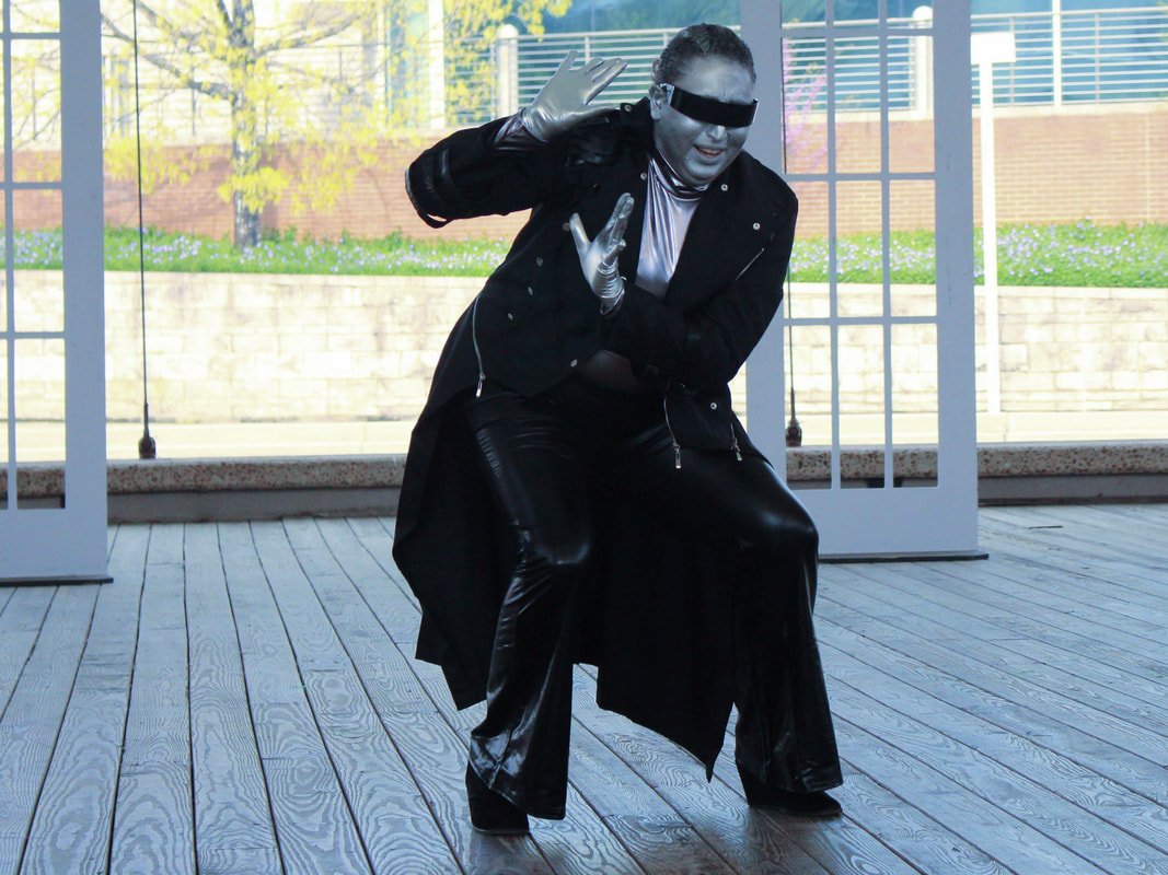 Person dressed as a robotic or futuristic character in black and silver attire, posing with arms raised.