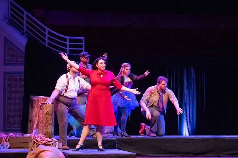 Actors performing on stage with colorful costumes, including a woman in a red dress and another in a blue tutu, with dramatic lighting and a dark curtain backdrop.