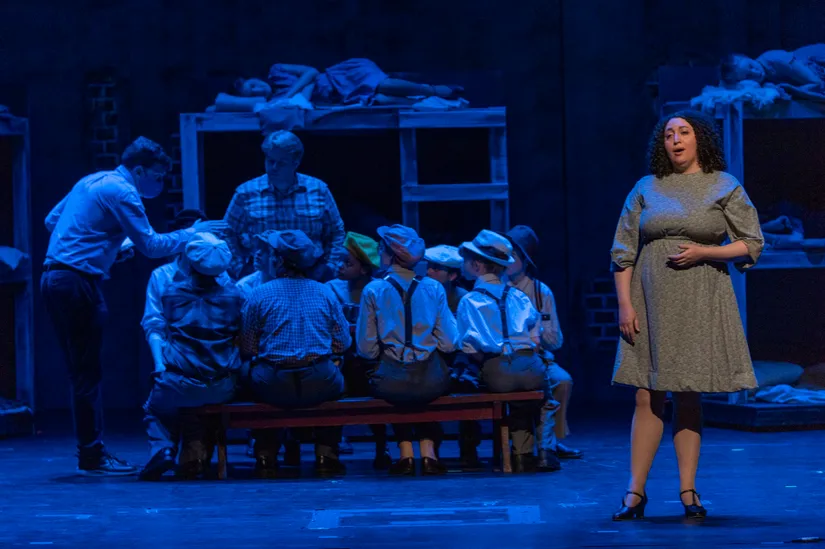 Stage performance with actors depicting a scene in a dormitory setting, with bunk beds and people gathered around a seated group. A woman in a dress is standing and singing or speaking.