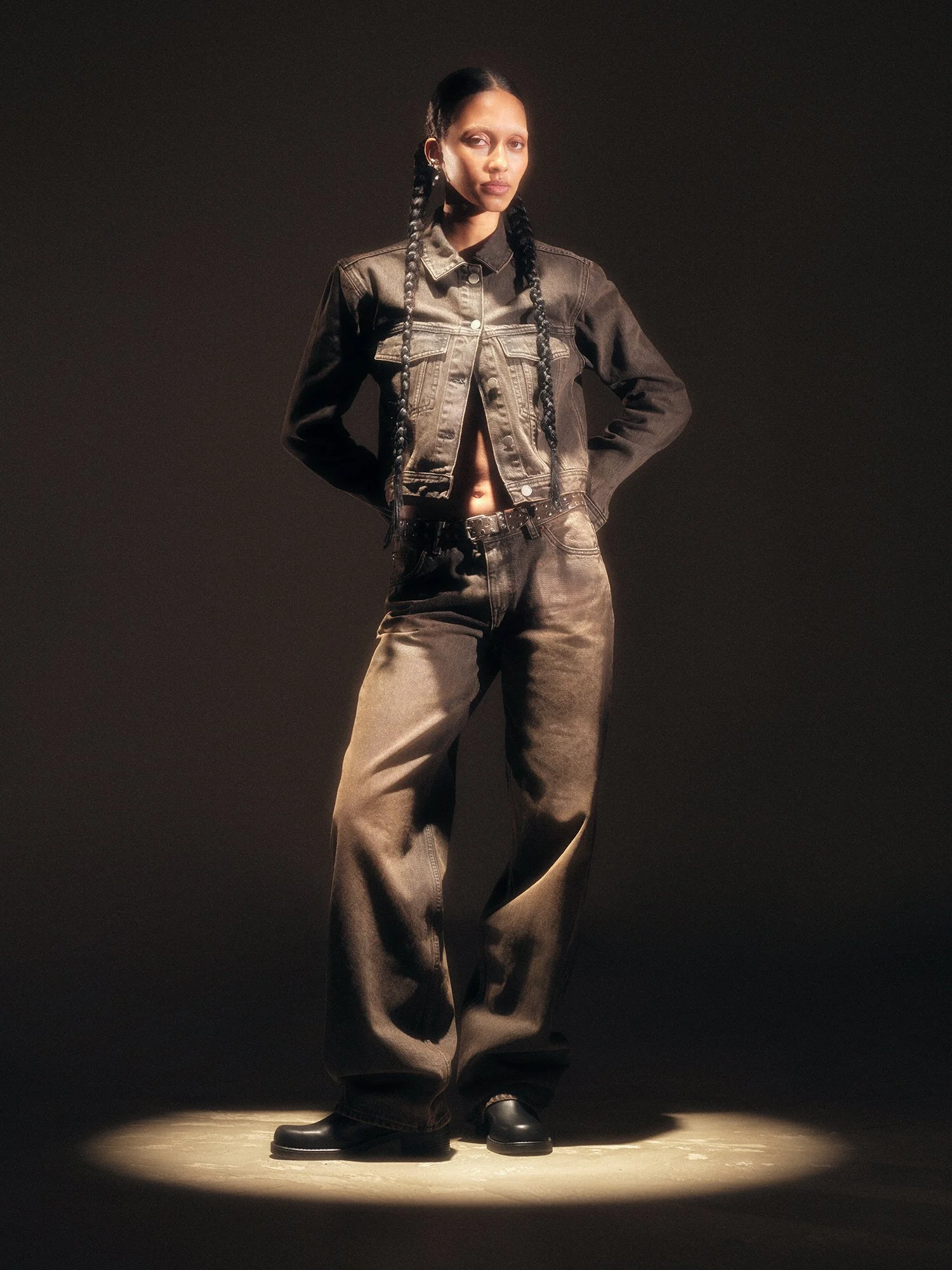 A woman standing on a spotlighted surface wearing a black denim jacket, black pants, and black boots, with her hands on her hips and her hair in two braids.