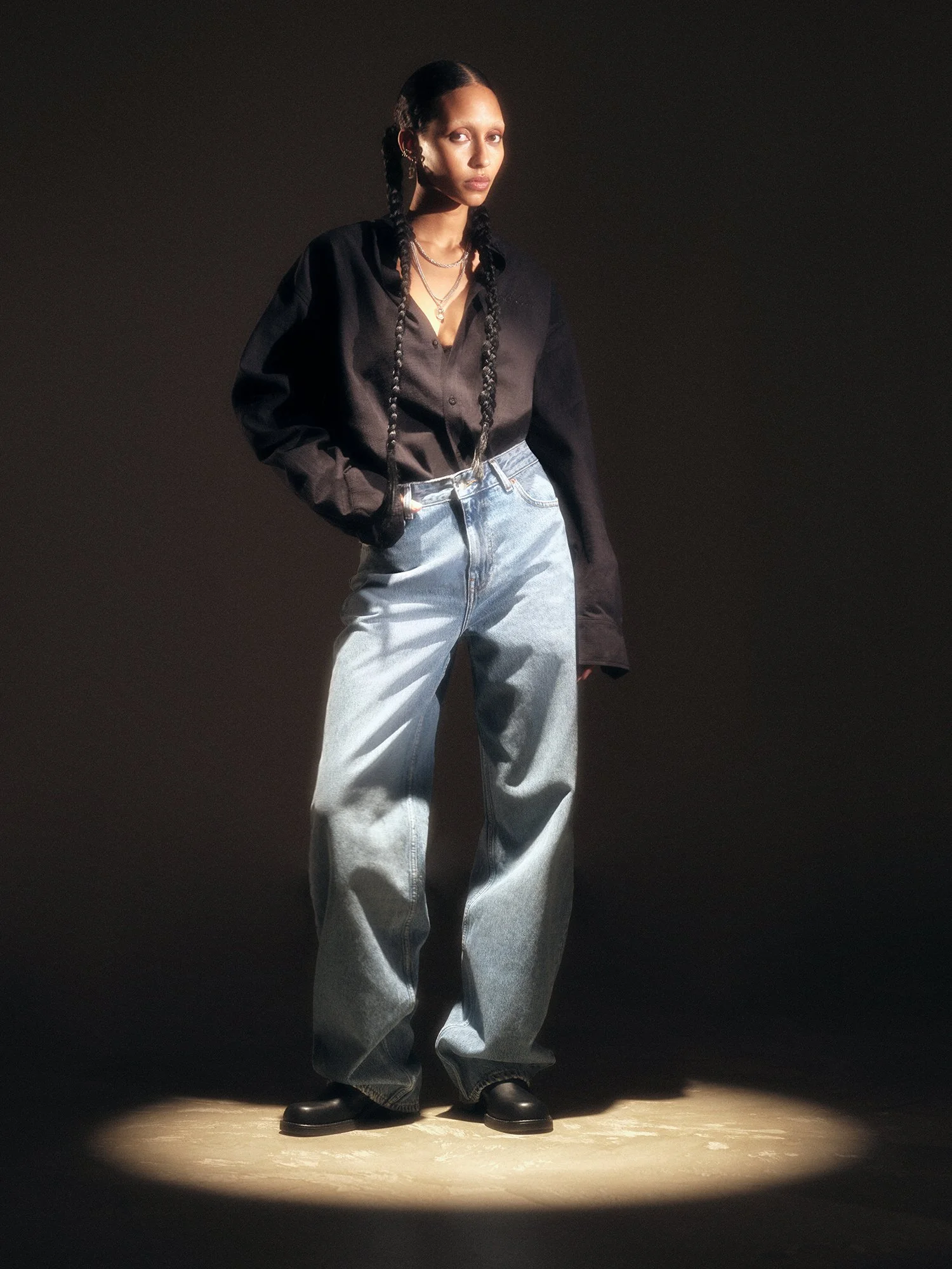 A woman standing in a spotlight wearing baggy light blue jeans, a black button-up shirt, and black shoes, with her hands in her pockets and long braided hair.