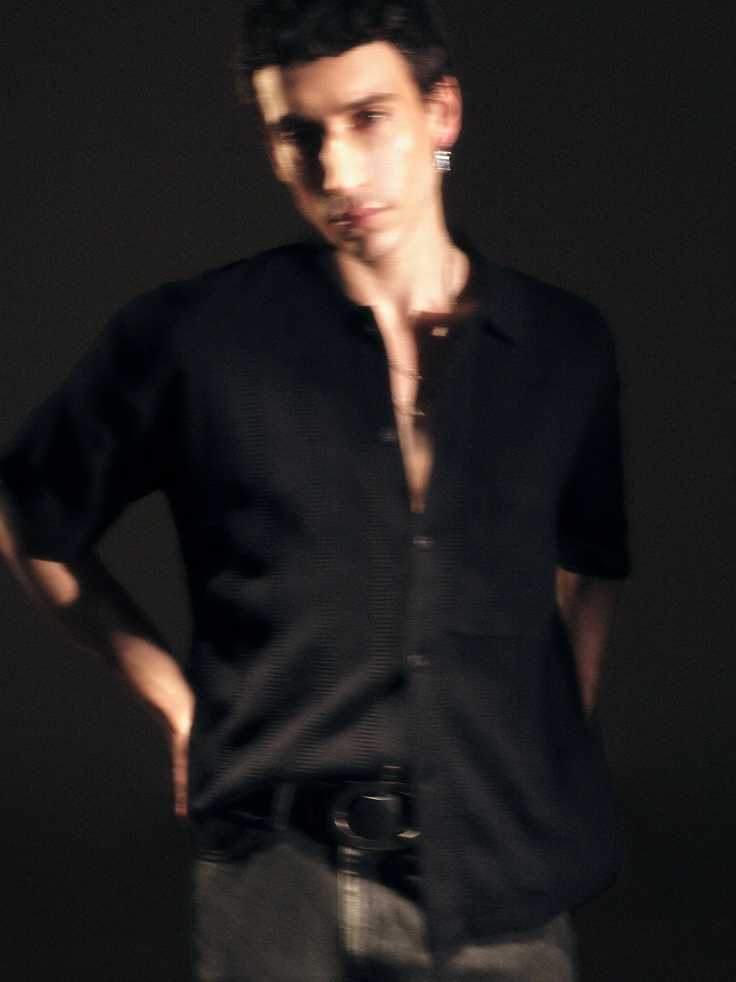 A blurred image of a young man with dark hair wearing a black shirt and a chain, standing against a dark background.
