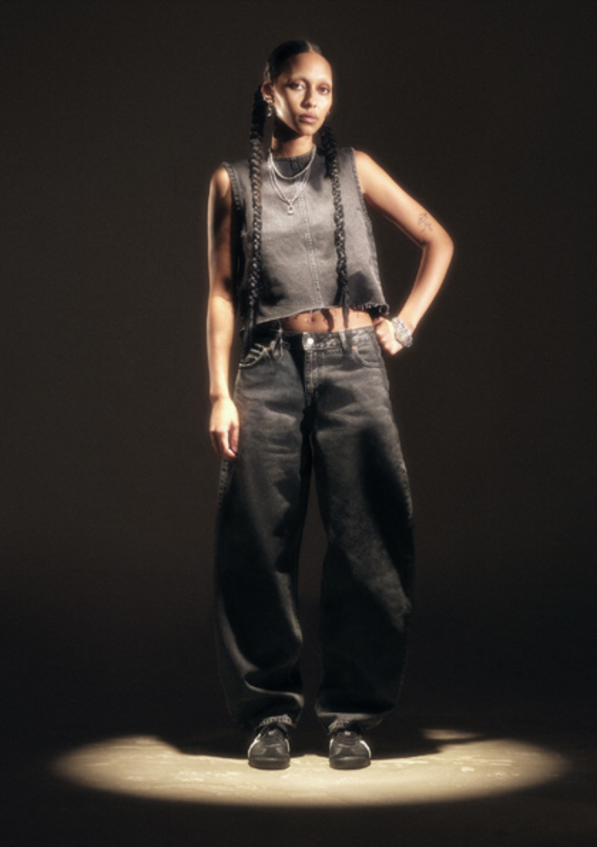 A woman standing solo in the spotlight against a dark background, dressed in casual streetwear, with long braids and jewelry.