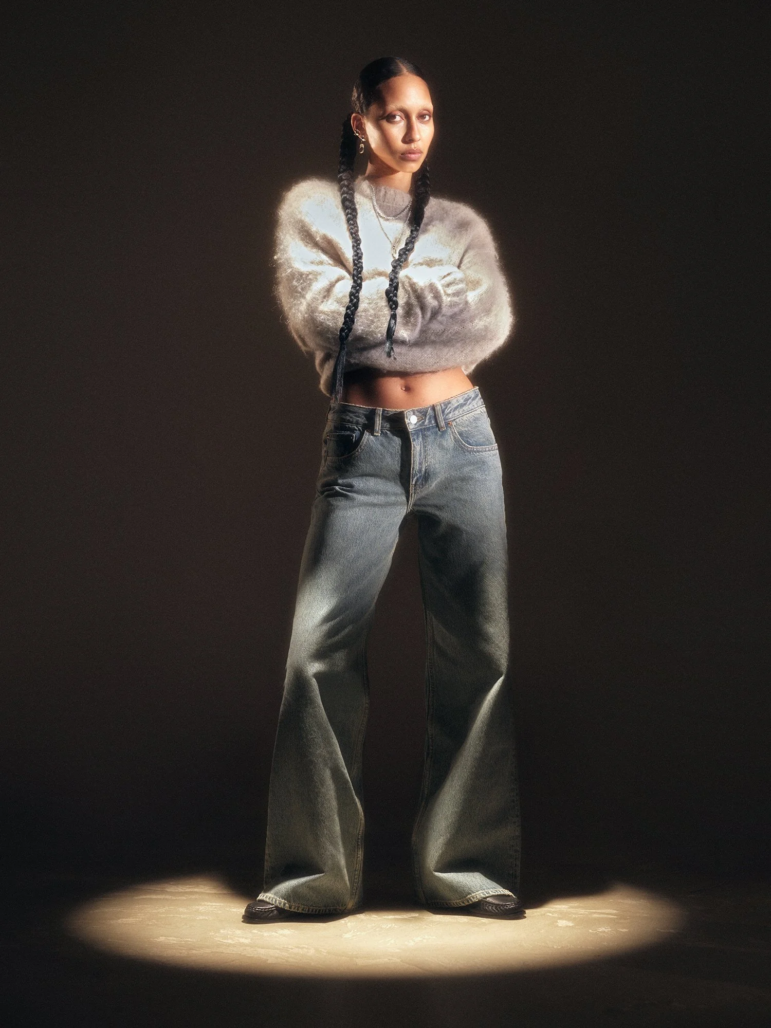 A woman with long braids stands in a spotlight with her arms crossed, wearing a fuzzy gray cropped sweater and baggy denim jeans.
