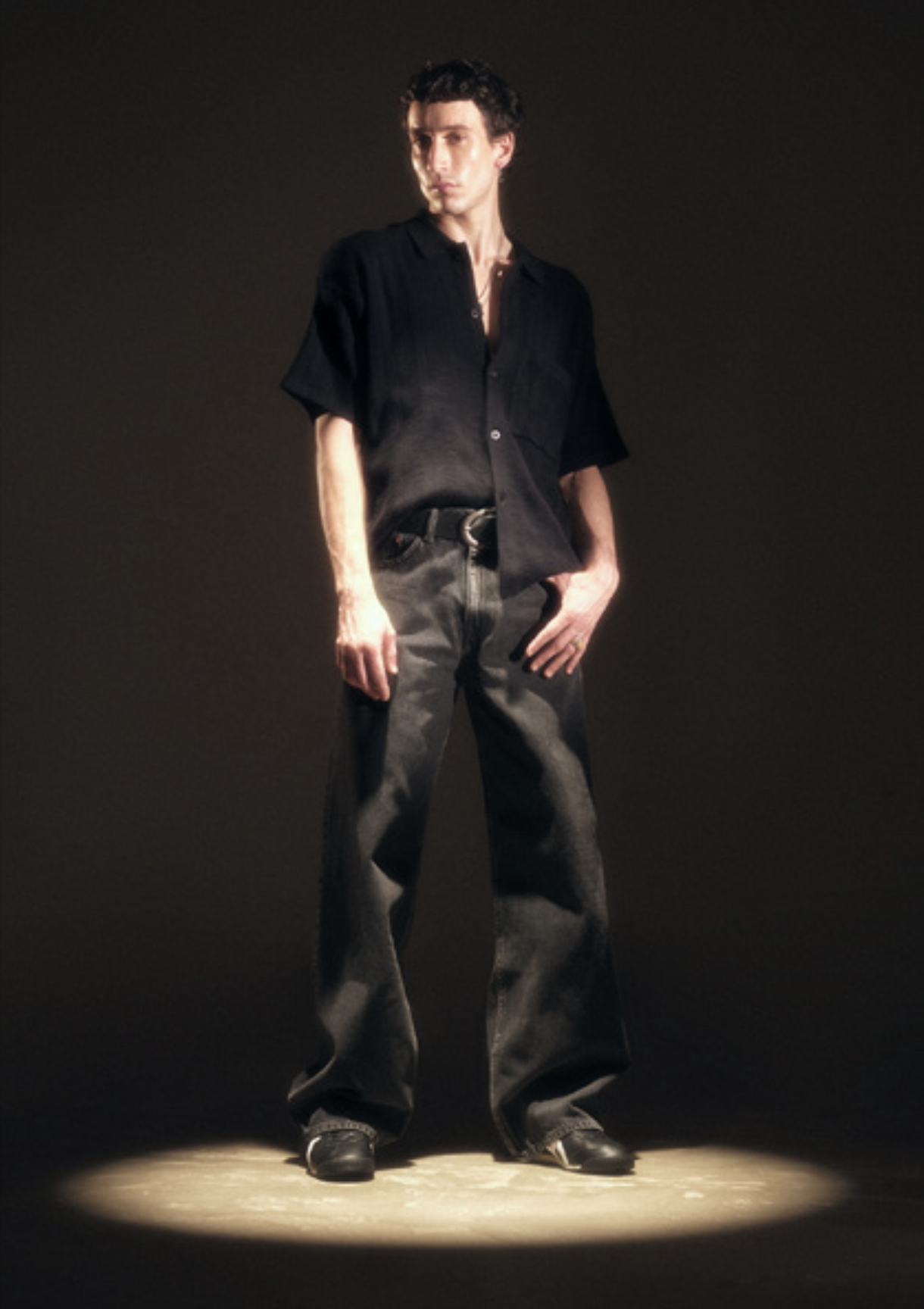 A young man standing in a spotlight with a dark background, wearing a black short-sleeved shirt, loose dark jeans, and cowboy boots.