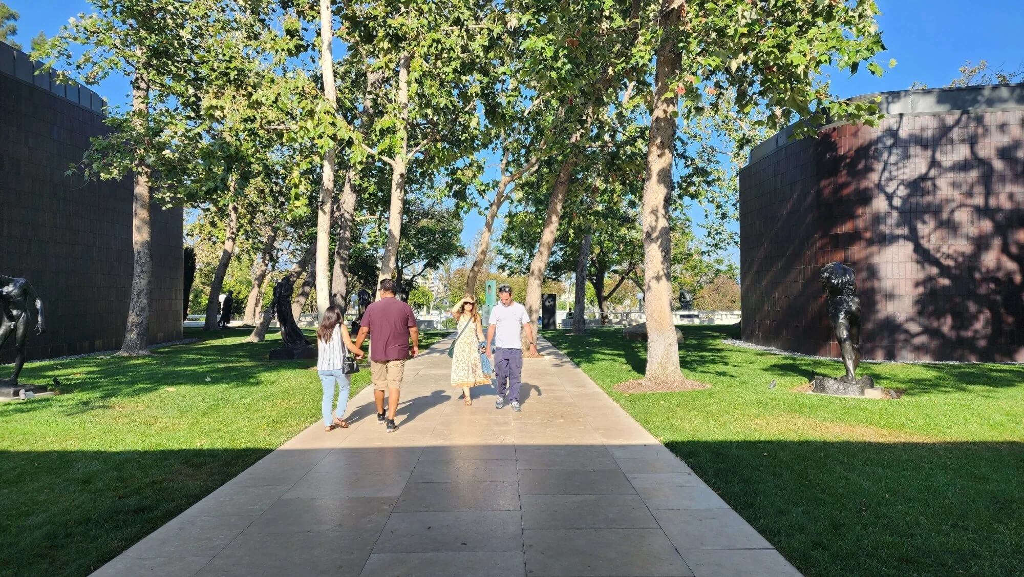 Tree-lined walkway outside the Norton Simon Museum in Pasadena, with bronze sculptures arranged across the lawn and visitors walking toward the museum.