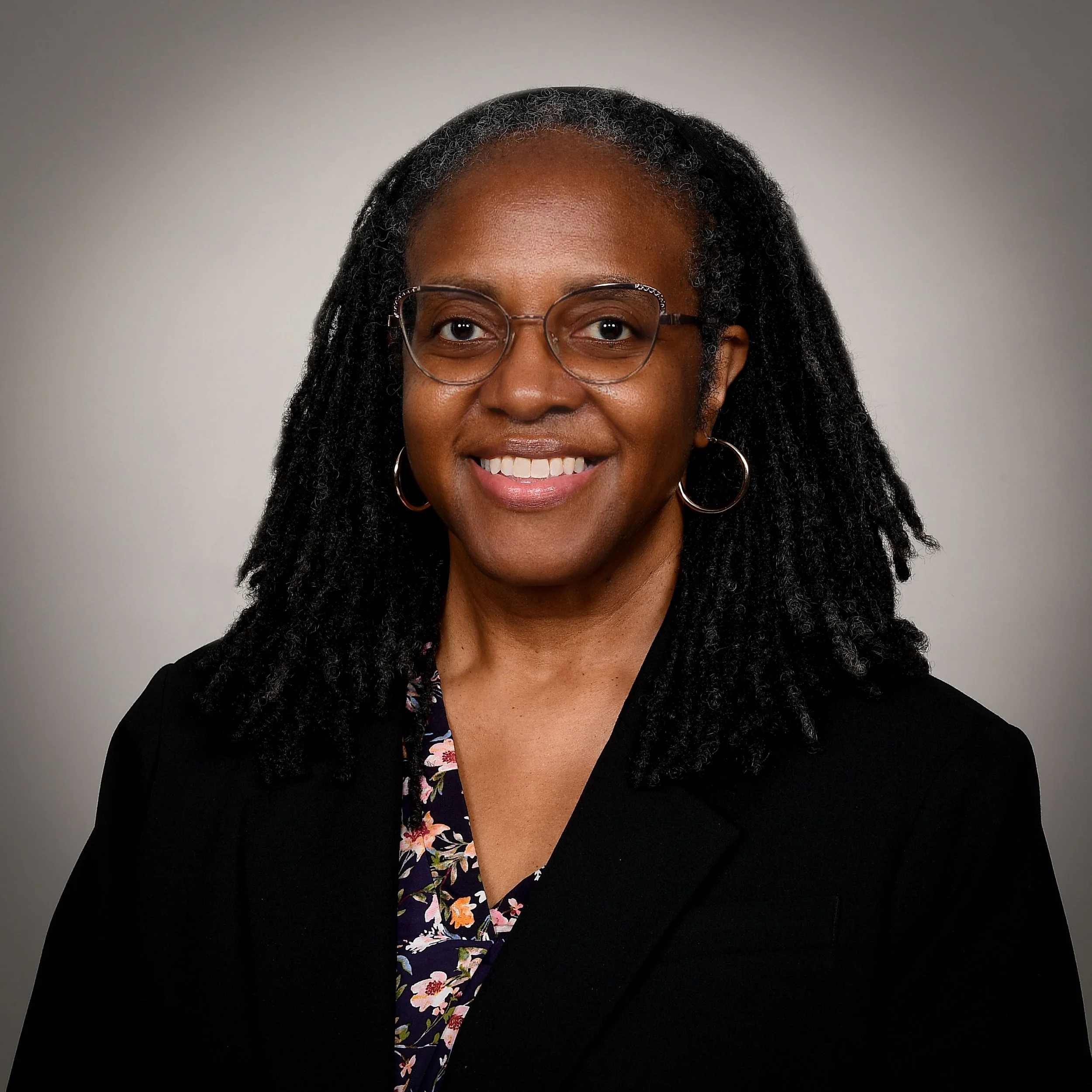 A smiling woman with glasses and hoop earrings, wearing a black blazer over a floral blouse, against a neutral grey background.