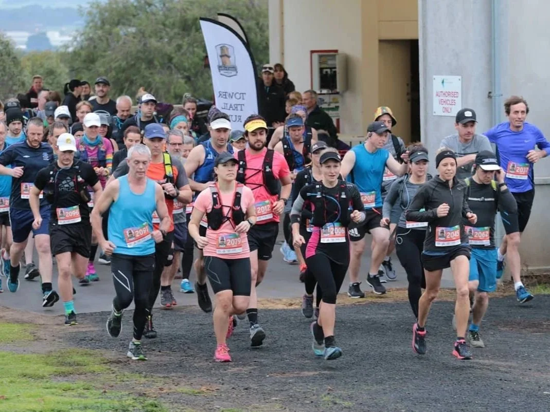 A group of runners participating in a marathon or race, running together on a paved path with some wearing bib numbers and athletic gear.