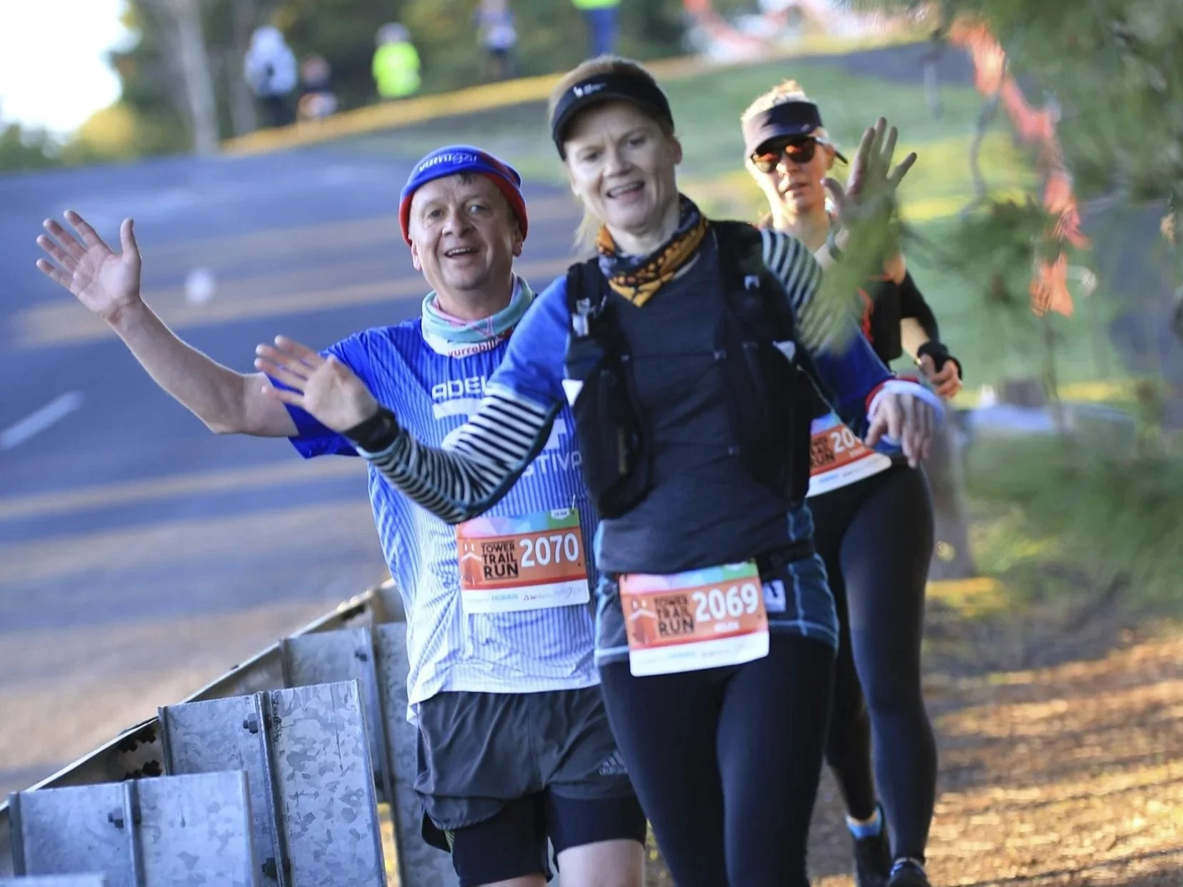 Group of runners participating in a trail race, smiling and waving at the camera, wearing race bibs and athletic gear, running along a wooded path.
