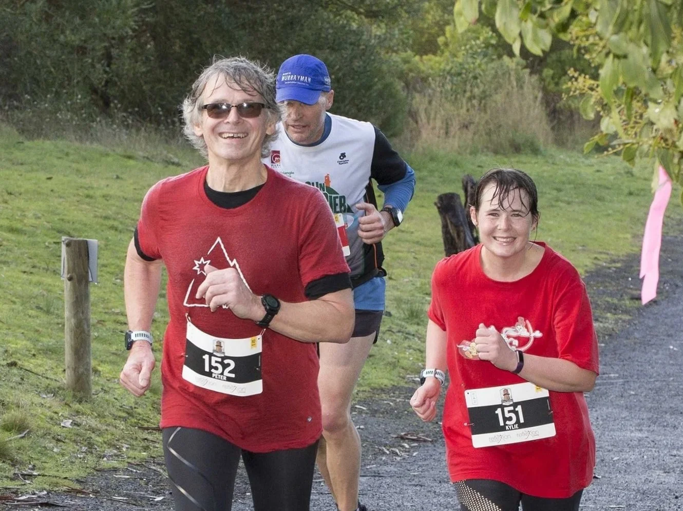 Three runners participating in a race on a trail, smiling and wearing race bibs, with trees and greenery in the background.