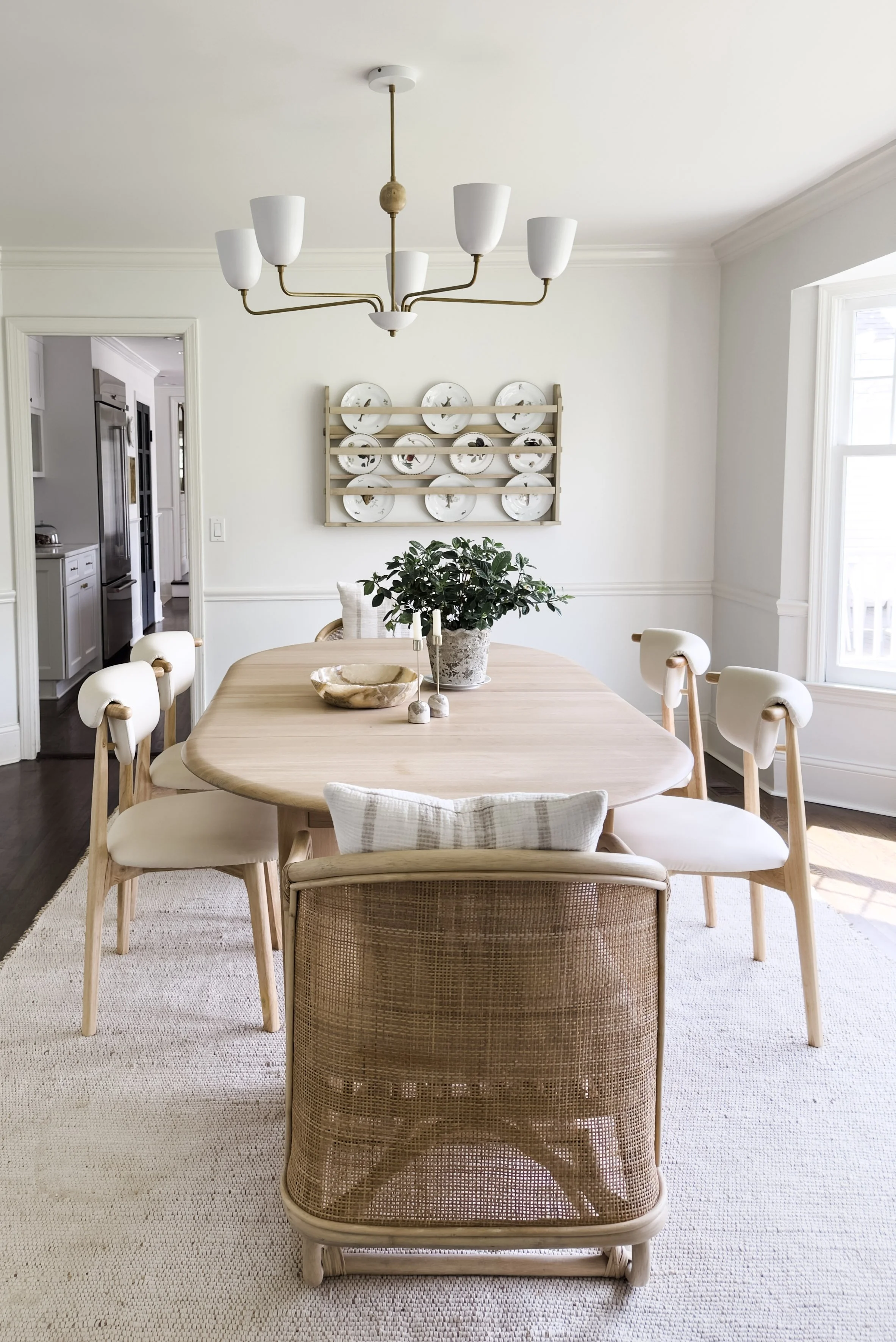 A dining room featuring a light wood oval table, surrounded by white and beige chairs. A large window on the right letting in natural light. A decorative wall hanging of plates on a wooden rack is on the far wall. A central chandelier with six white 