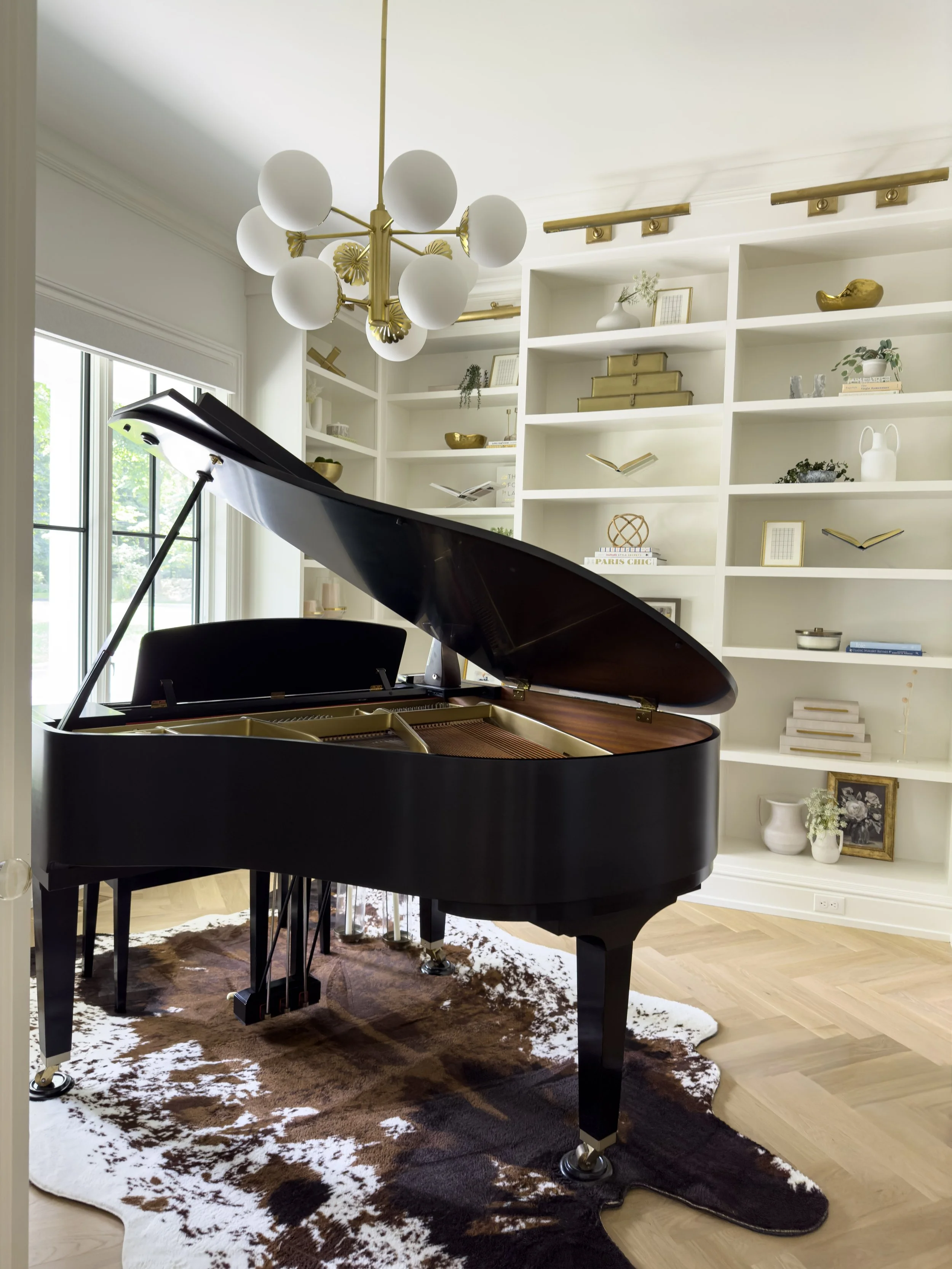 A black grand piano on a cowhide rug in a bright room with built-in white shelves decorated with books, vases, and artwork, and a large window with greenery outside.