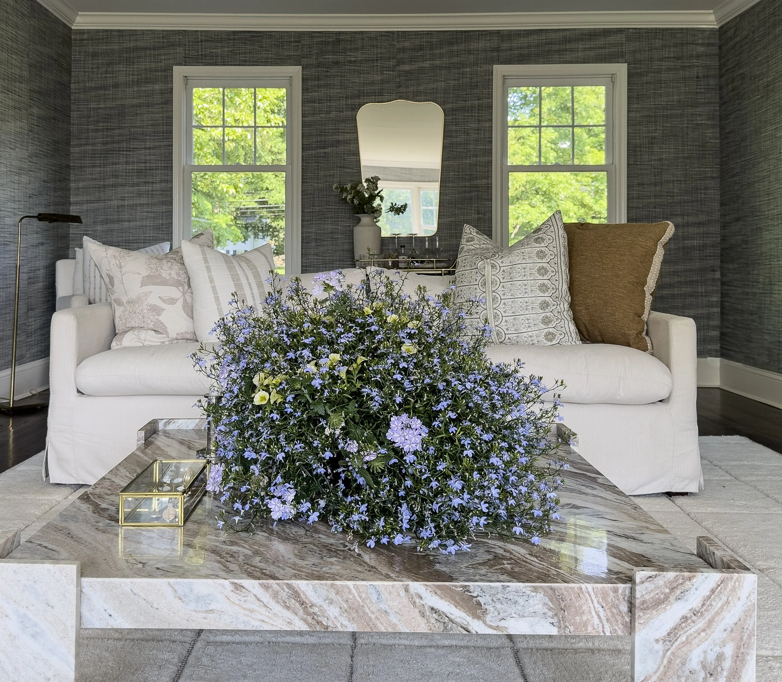 Living room with two large windows showing green trees outside, a white sofa with patterned pillows, a glass coffee table with a gold frame and a bouquet of blue and white flowers, a mirror, a vase with flowers, a brass tray, and a marble hearth.