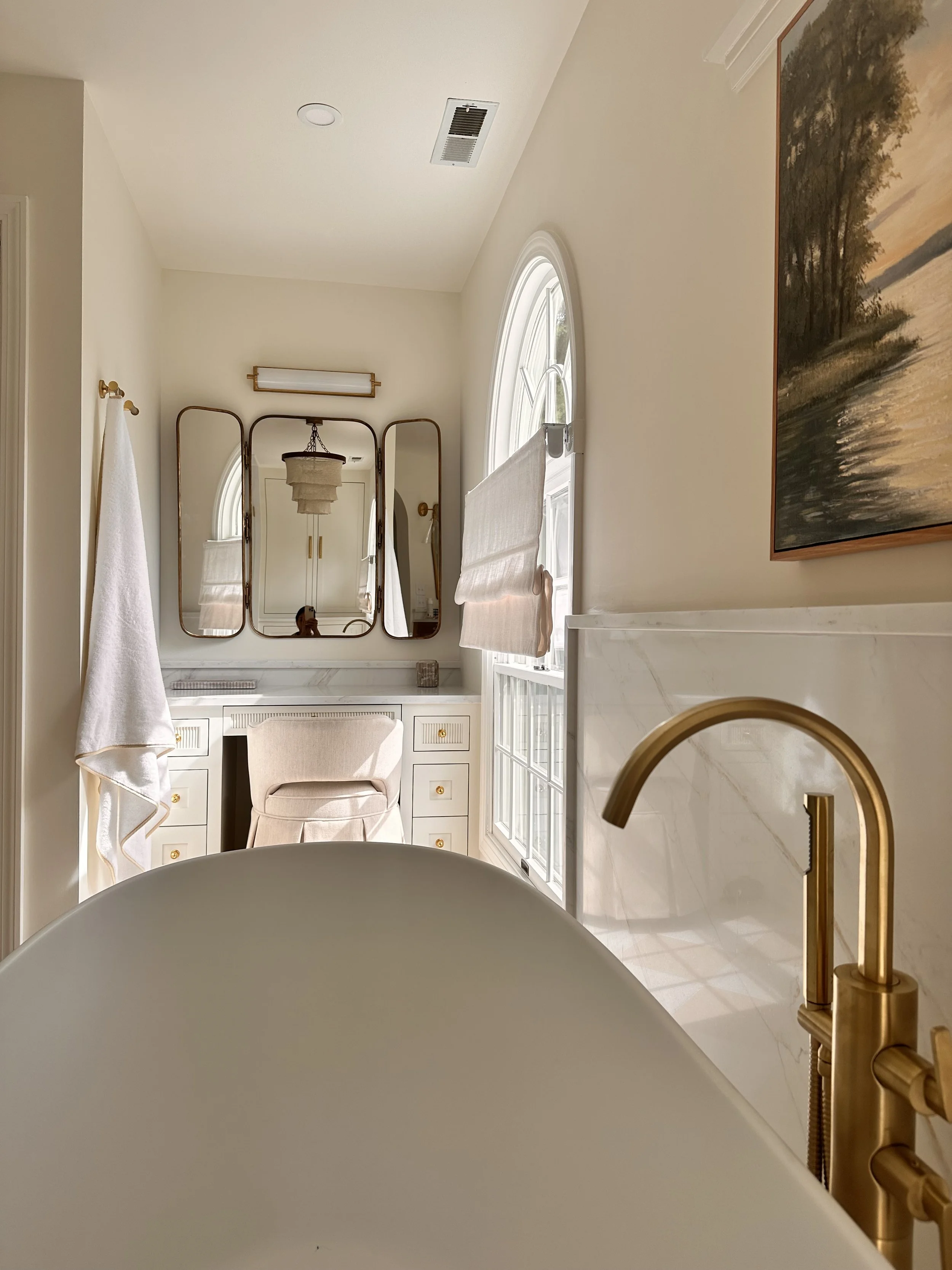 Close-up view of a modern bathroom sink with a gold faucet, a bathtub in the foreground, a window with curtains, a white chair, a mirror, and a painting of a landscape scene on the wall.