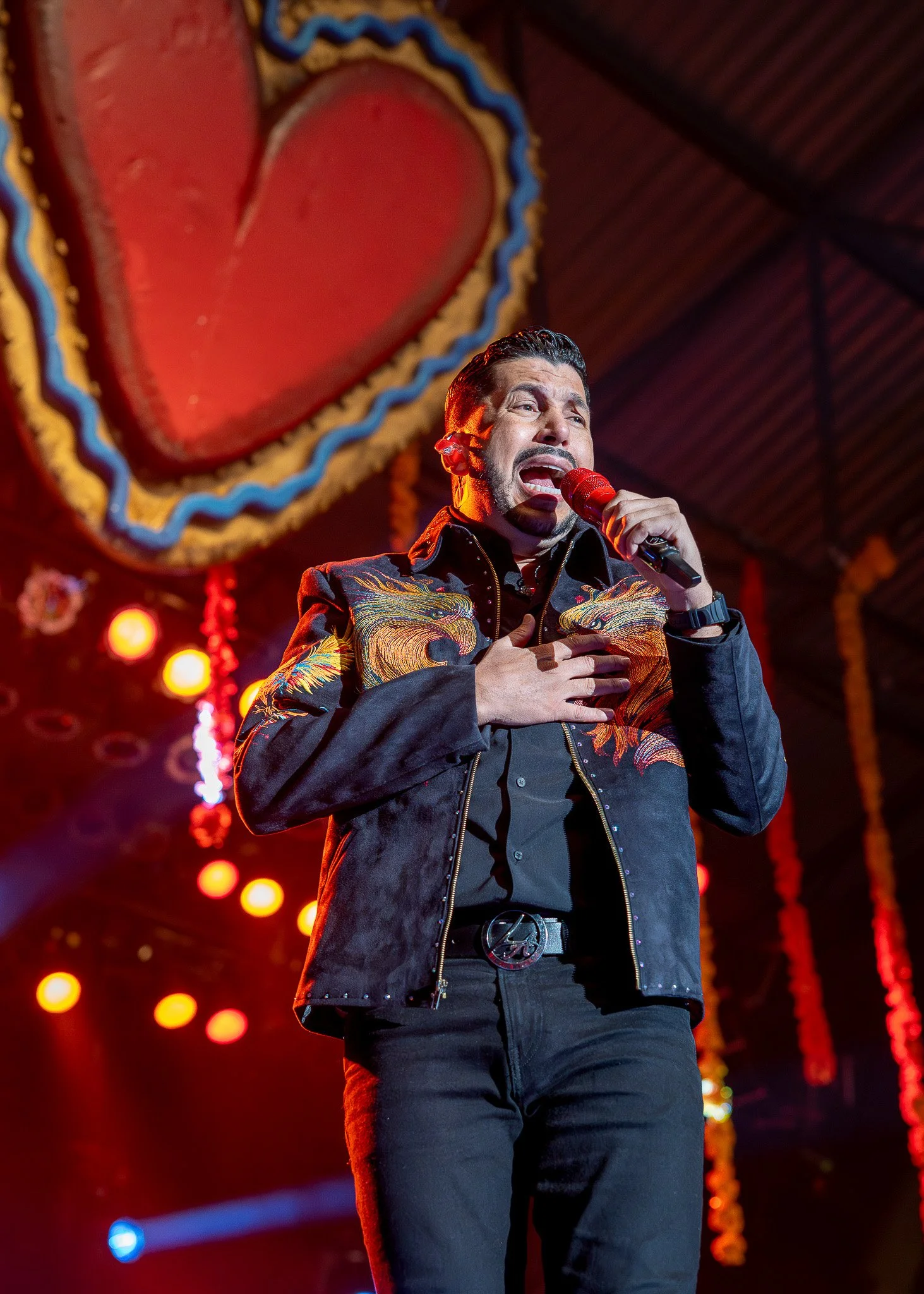 A singer performing on stage, holding a red microphone with his right hand and placing his left hand on his chest. He is wearing a black jacket with colorful embroidered dragons and a belt with a peace sign buckle. The background features colorful circular stage lights and red decorative hanging ornaments. There is a large, illuminated red heart-shaped decoration with a blue border hanging above him.