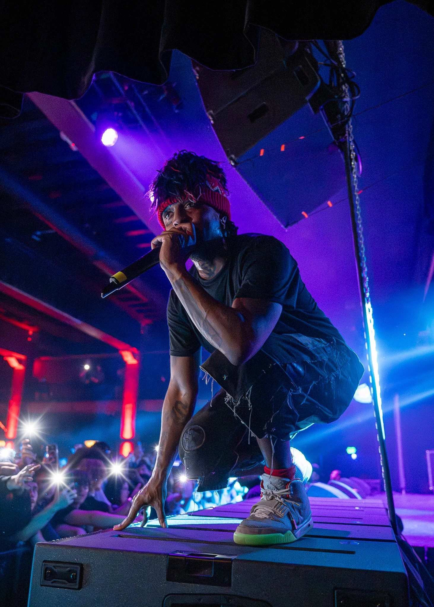 Performer crouching on stage with microphone in hand at a concert, audience taking photos, colorful stage lighting, and speakers in the background.