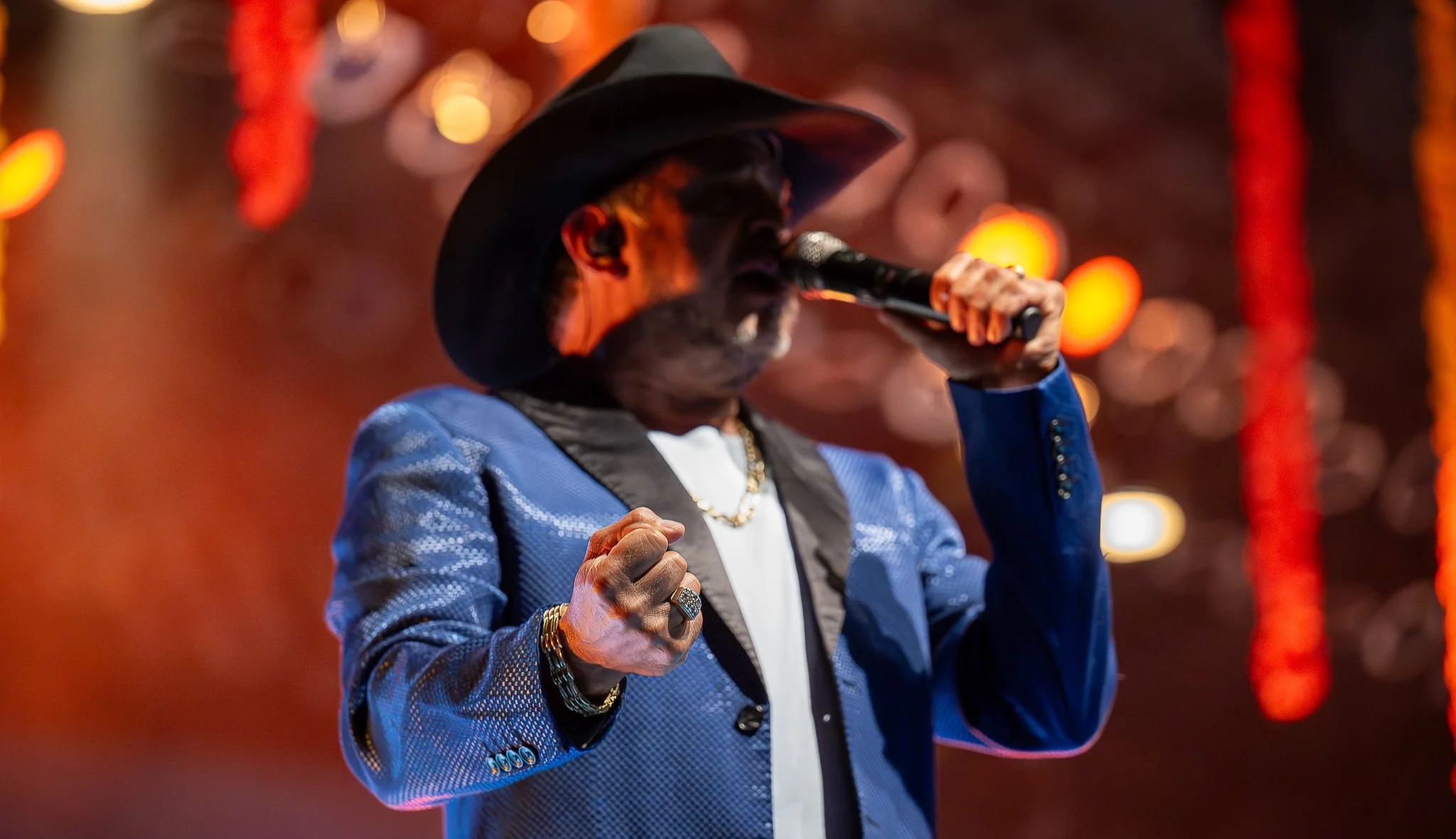 A man wearing a black cowboy hat and a shiny blue jacket singing into a microphone on stage, with orange and red lights in the background.