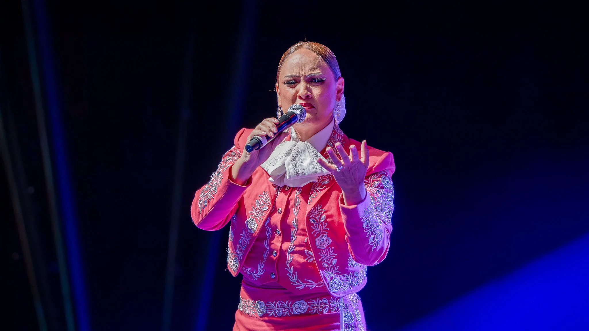 A woman singing on stage, wearing a pink embroidered outfit with a white lace collar, holding a microphone, with a serious expression.