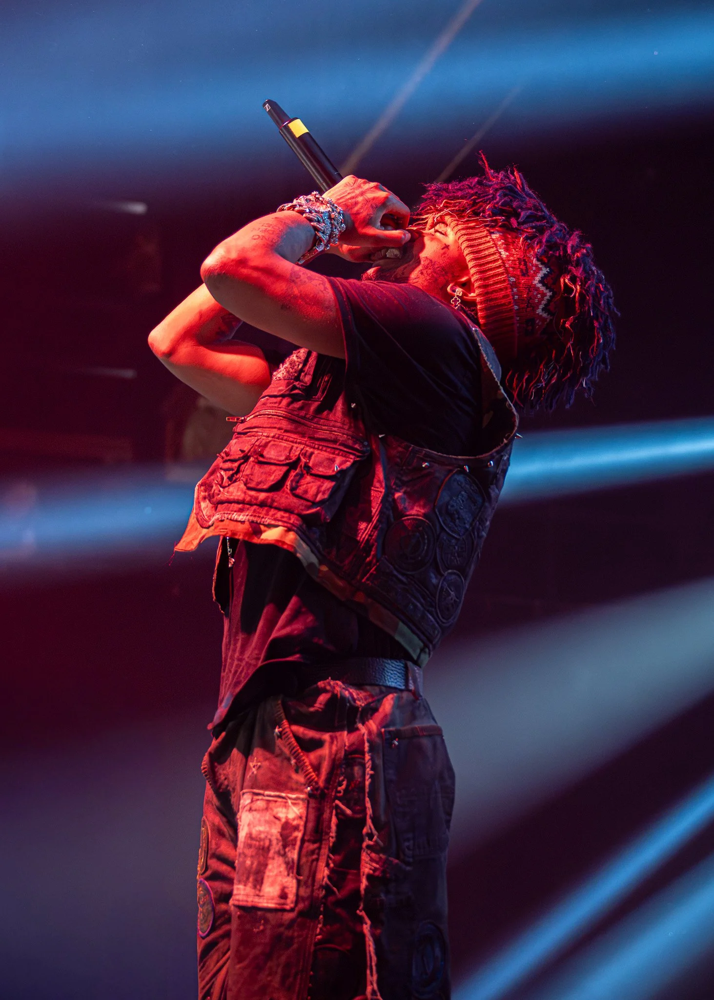 A male performer with dreadlocks, wearing a colorful hat and a black vest, singing into a microphone on stage under colorful lights.
