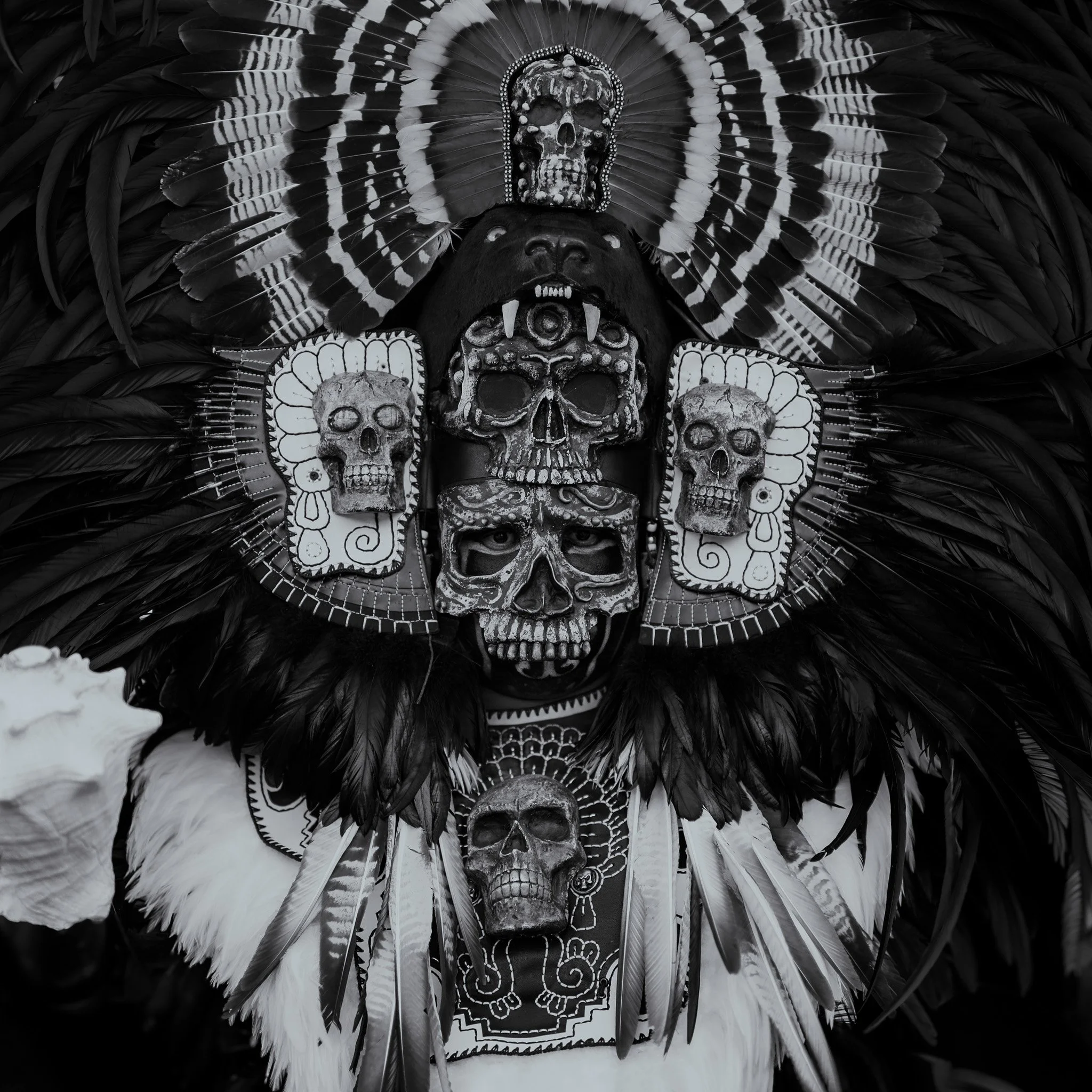 A person wearing a traditional indigenous ceremonial headdress decorated with skulls, feathers, and intricate patterns, with face painted and skull masks.
