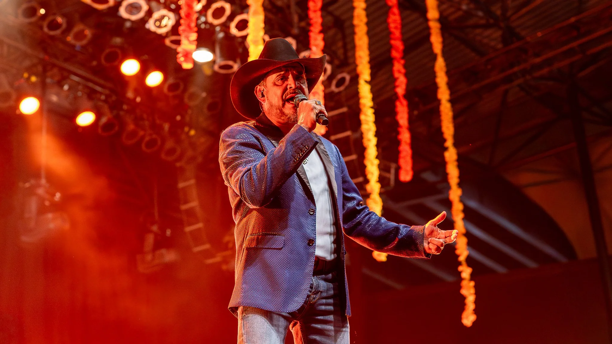 A male performer singing into a microphone on stage, wearing a cowboy hat and a blue blazer, with orange stage lights and hanging decorations in the background.