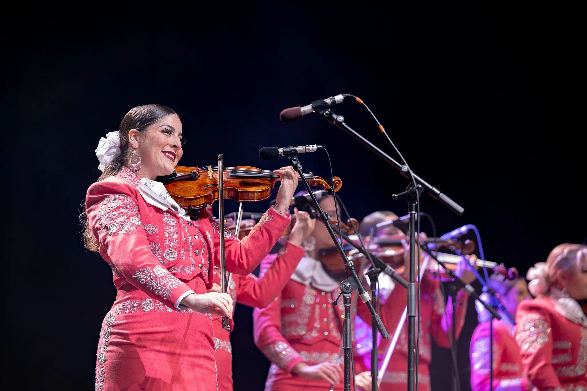 A woman in a pink embroidered dress playing a violin during a performance with multiple microphone stands on stage.